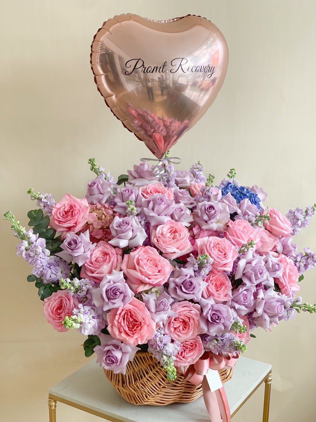 A luxurious floral basket arrangement featuring a blend of pink and lavender roses, delicate stock flowers, and lush greenery, topped with a heart-shaped 