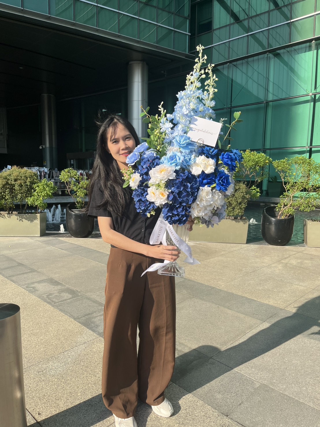 A woman stands outdoors in sunlight, holding a large bouquet of artificial flowers in blue tone with a card attached. She wears a black shirt and brown trousers, surrounded by green plants and a glass building in the background.