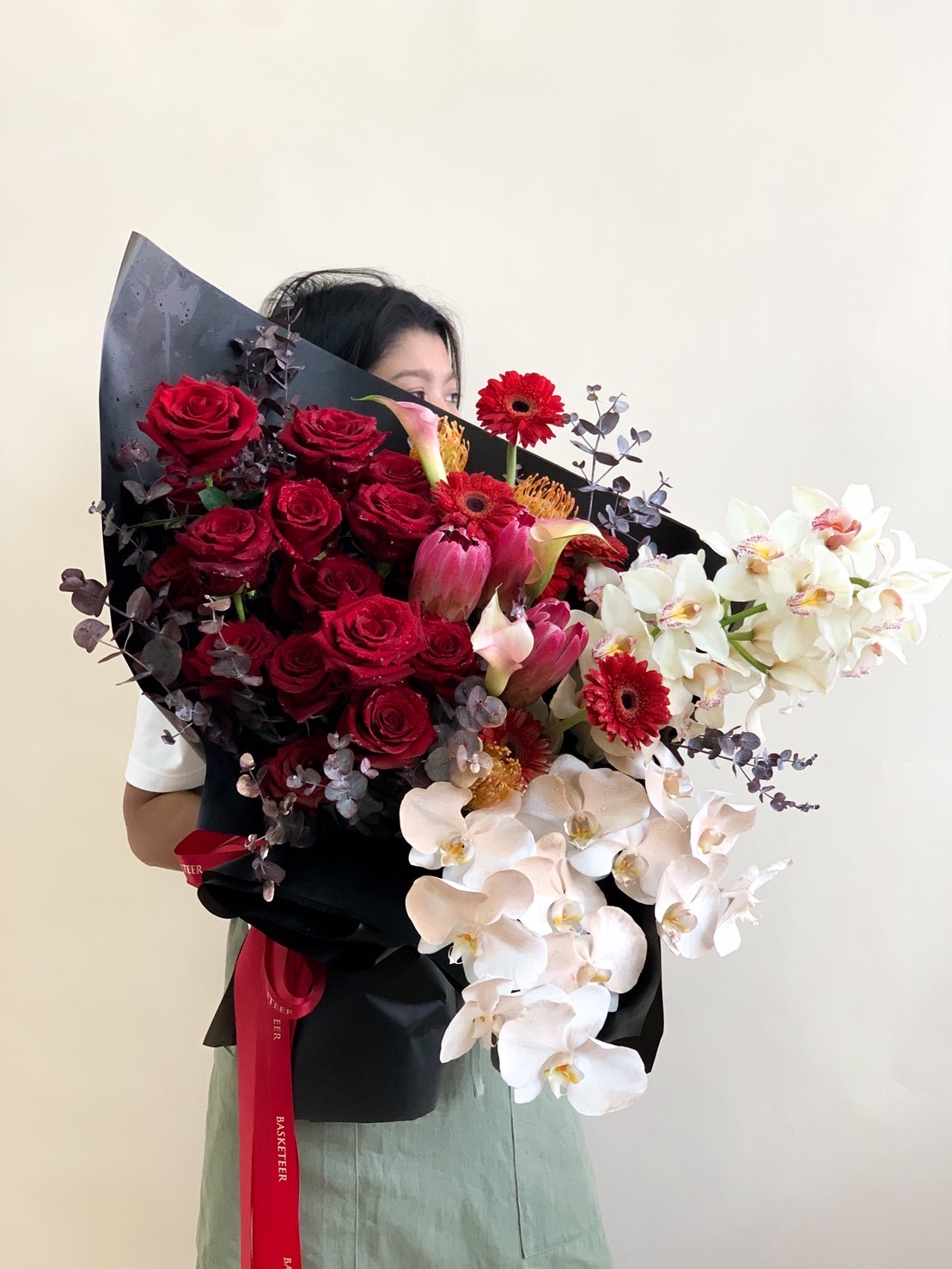 Large luxury bouquet with red roses, white orchids, red gerberas, protea, and dark eucalyptus wrapped in black paper.