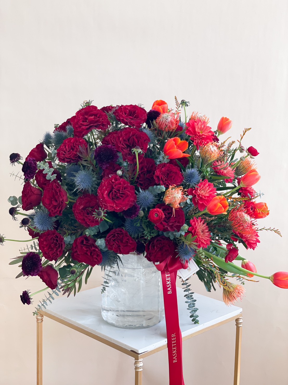 A luxurious red and orange floral arrangement featuring roses, tulips, and exotic flowers in a glass vase with a red ribbon.