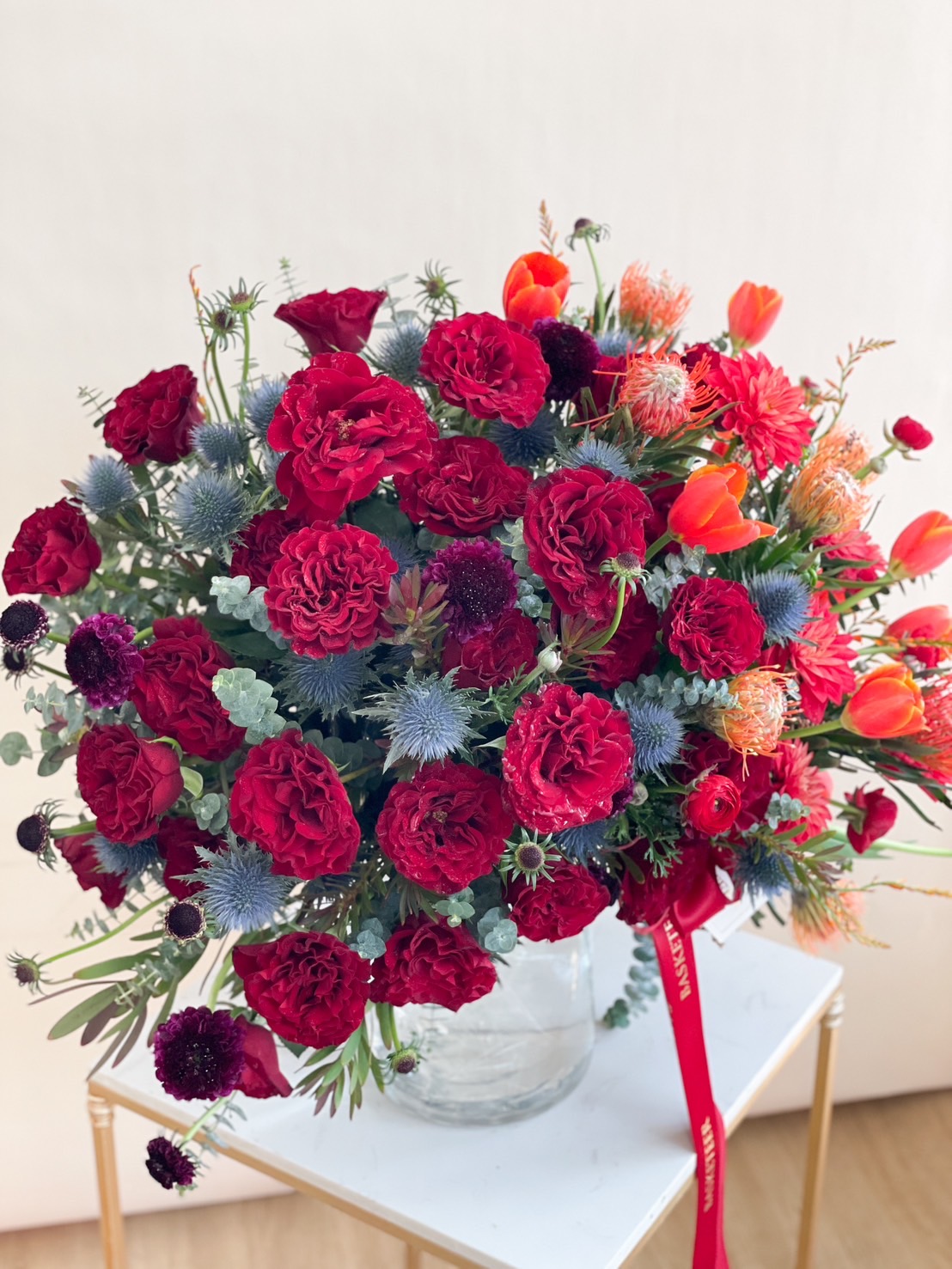 A luxurious red and orange floral arrangement featuring roses, tulips, and exotic flowers in a glass vase with a red ribbon.