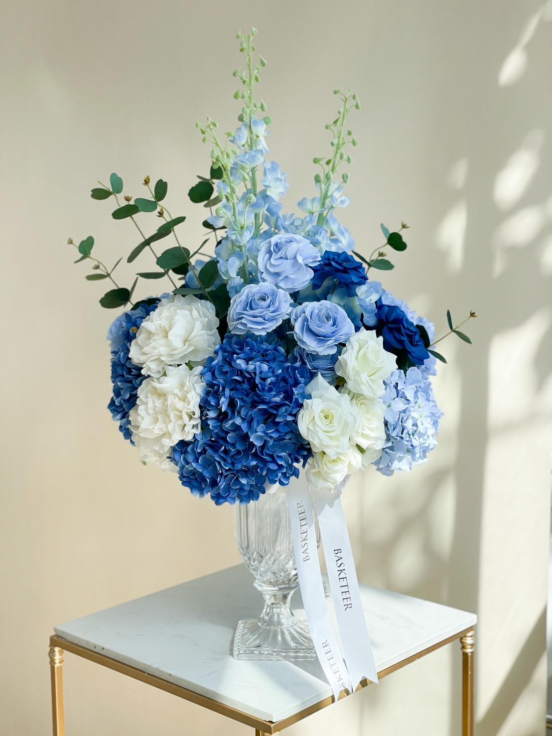 A bouquet of artificial blue and white flowers arranged in a clear glass vase on a gold and white pedestal table. The bouquet includes roses, hydrangeas, and delphiniums, with green foliage accents. Two white ribbons are attached, with 