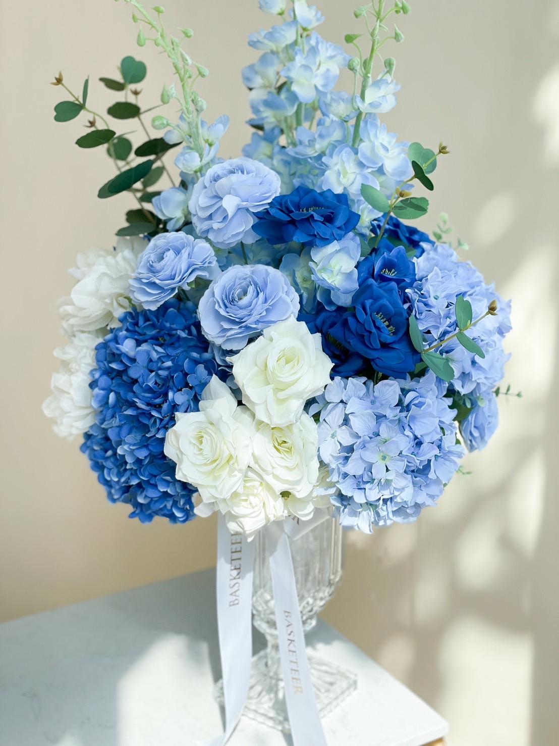 A vibrant bouquet in a glass vase, featuring an array of blue and white blooms including hydrangeas, roses, and lisianthus. Eucalyptus leaves add a touch of greenery. The vase with artificial flowers in blue tones has white ribbons attached, reading 