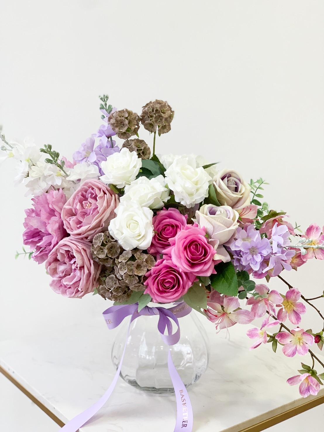 A romantic floral arrangement in a clear glass vase features a mix of pink, white, and lavender roses, alongside light purple flowers and green foliage. A light purple ribbon is tied around the vase, accentuating its charming purple tone. The background is a plain off-white color.