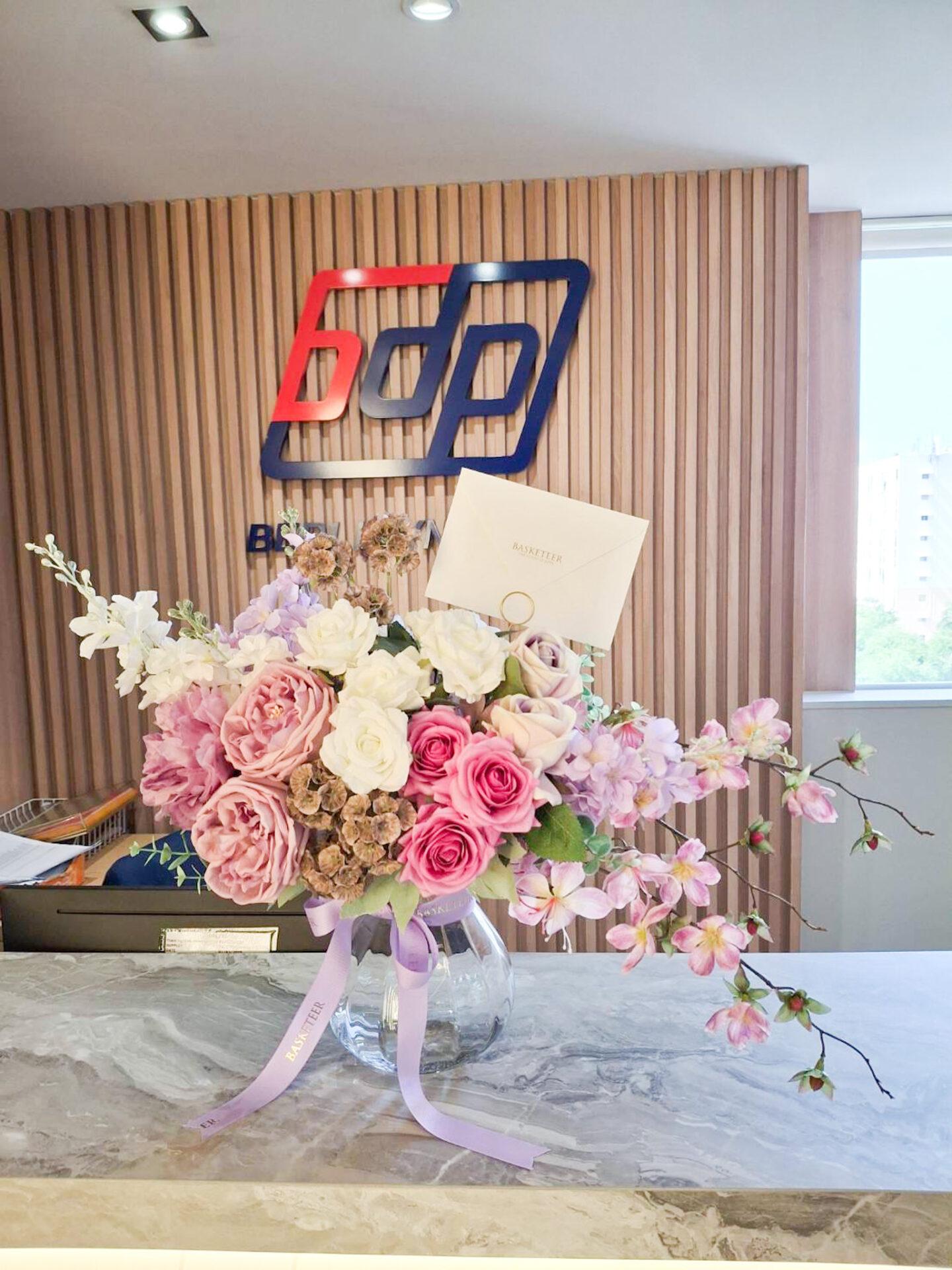 A glass vase filled with an arrangement of romantic artificial flowers in pink, white, and purple tones is placed on a marble counter in front of a wooden backdrop. A logo featuring the letters 