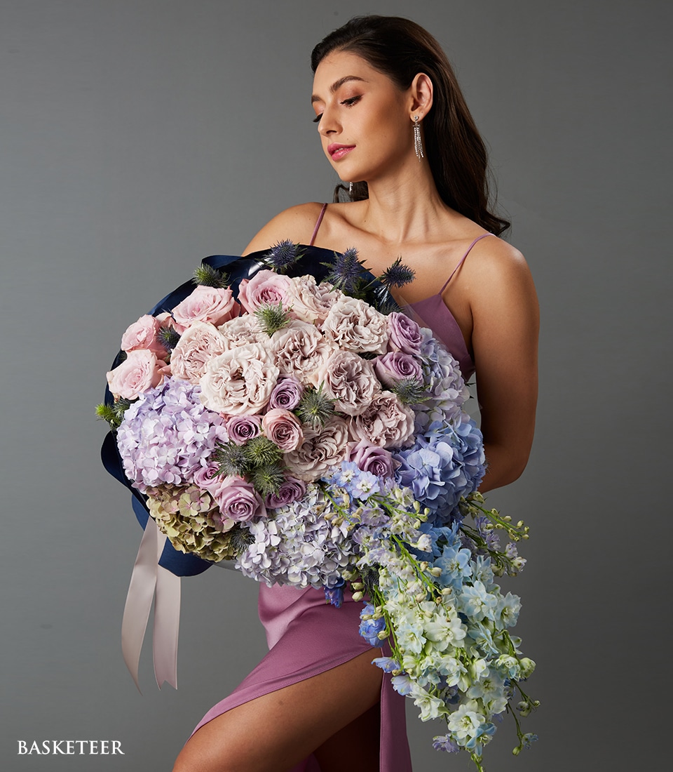 A woman in an elegant lavender dress cradles a sumptuous, oversized bouquet of flowers. The exquisite arrangement showcases a harmonious blend of pastel-colored roses and an assortment of enchanting purple and blue blooms. She gazes downward with a tranquil expression, capturing the essence of serene beauty against a sophisticated gray backdrop. In the corner, 