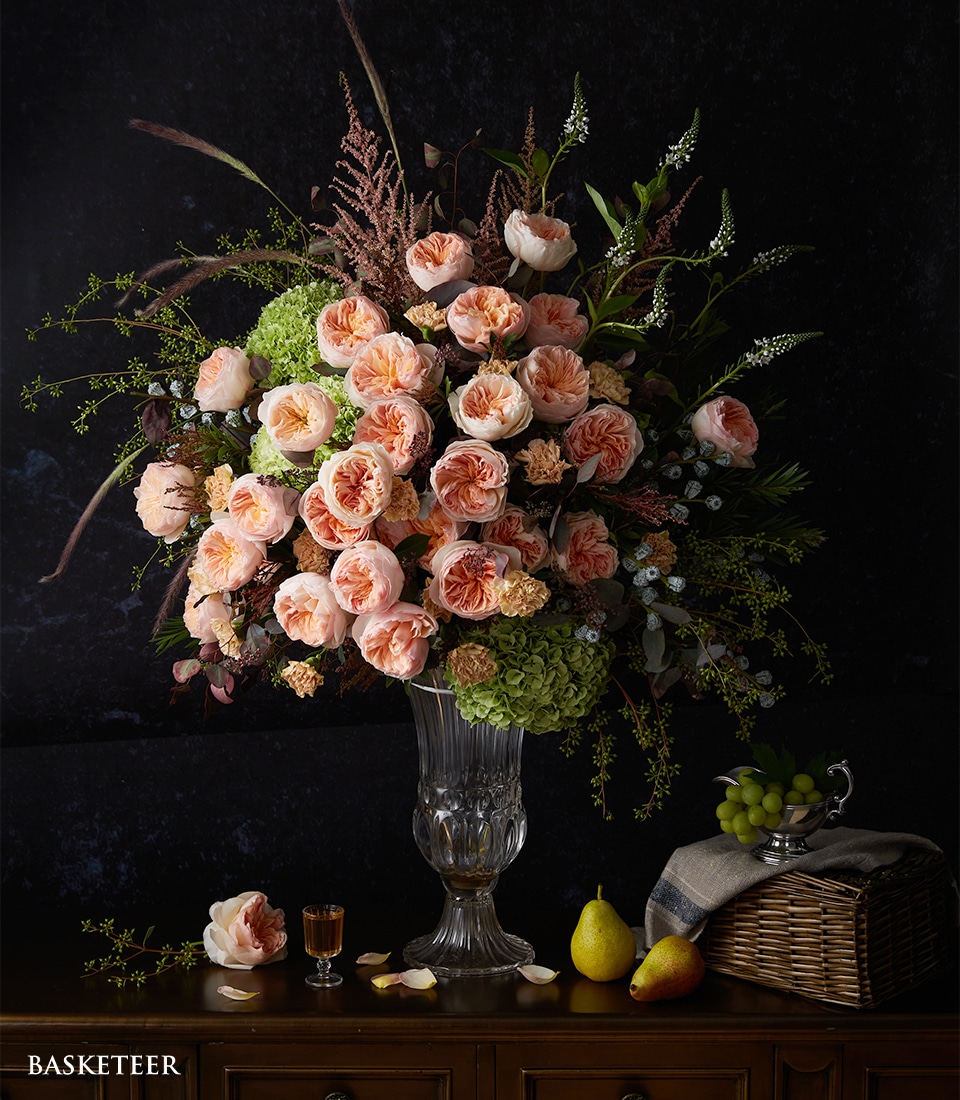 A lavish bouquet of pink and peach roses, lush green hydrangeas, and verdant foliage arranged elegantly in a crystal-clear glass vase graces a rich wooden surface. Enhancing the opulence, a wicker basket brimming with ripe grapes sits nearby. A refined glass of amber liquid complements the arrangement alongside a succulent pear. Delicate petals are artfully scattered around, adding to this luxurious tableau. Notice the masterful touch – a ribbon adorned with the distinguished Basketeer logo draping subtly over the vase, promising an extraordinary experience from Basketeer.