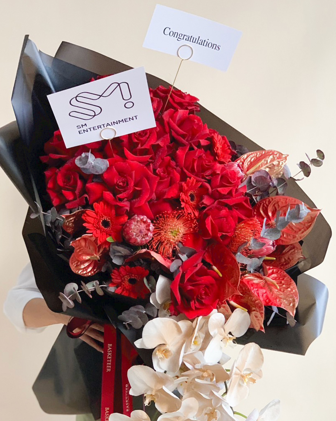 A person holds a Luxurious Scarlet Flowers Bouquet of red and white blooms wrapped in black paper, featuring a 