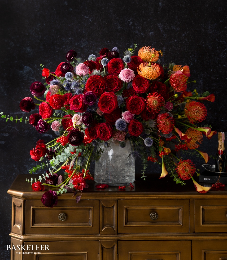 A sumptuous arrangement of vibrant flowers, featuring elegant red roses, striking orange pincushion proteas, and charming pink globular blooms, graces a clear vase positioned on a finely crafted wooden dresser. A bottle of exquisite champagne stands beside it. The distinguished 