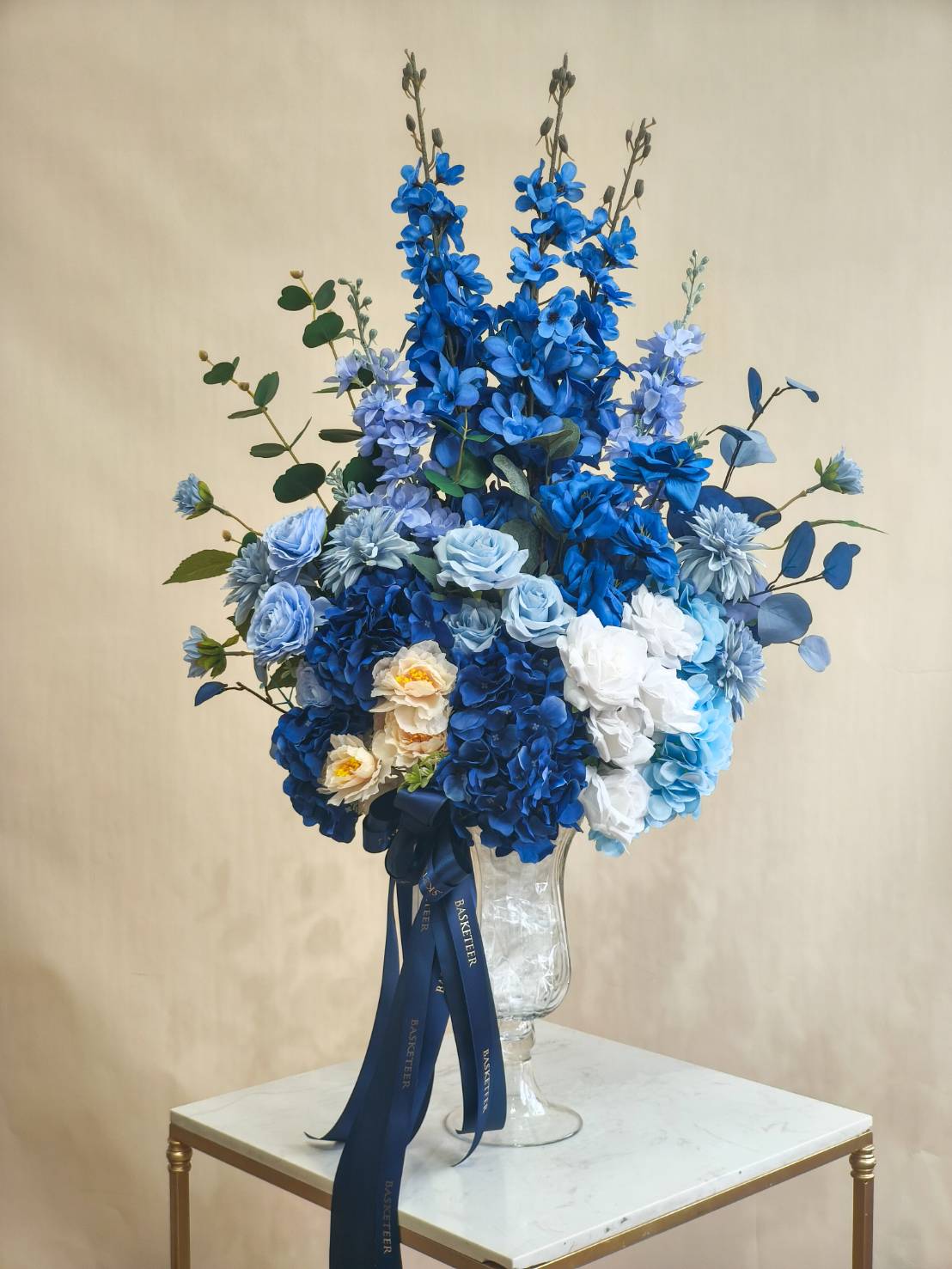Luxury tall flower vase with blue delphiniums, hydrangeas, roses, and white peonies, accented with greenery and tied with a navy satin ribbon.