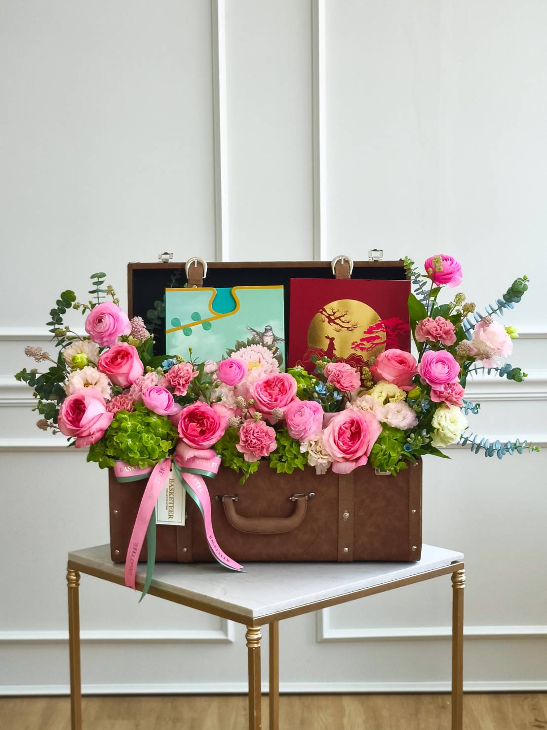 Luxury leather suitcase flower arrangement with pink roses, ranunculus, carnations, and gift boxes for premium celebrations