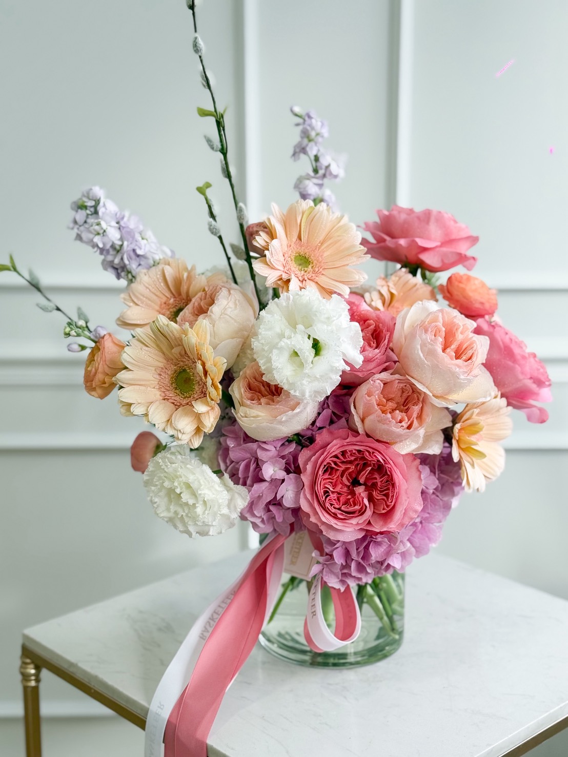 Elegant pastel flower vase with pink peonies, gerbera, ranunculus, hydrangeas, lisianthus, and stock in a glass vase with ribbon