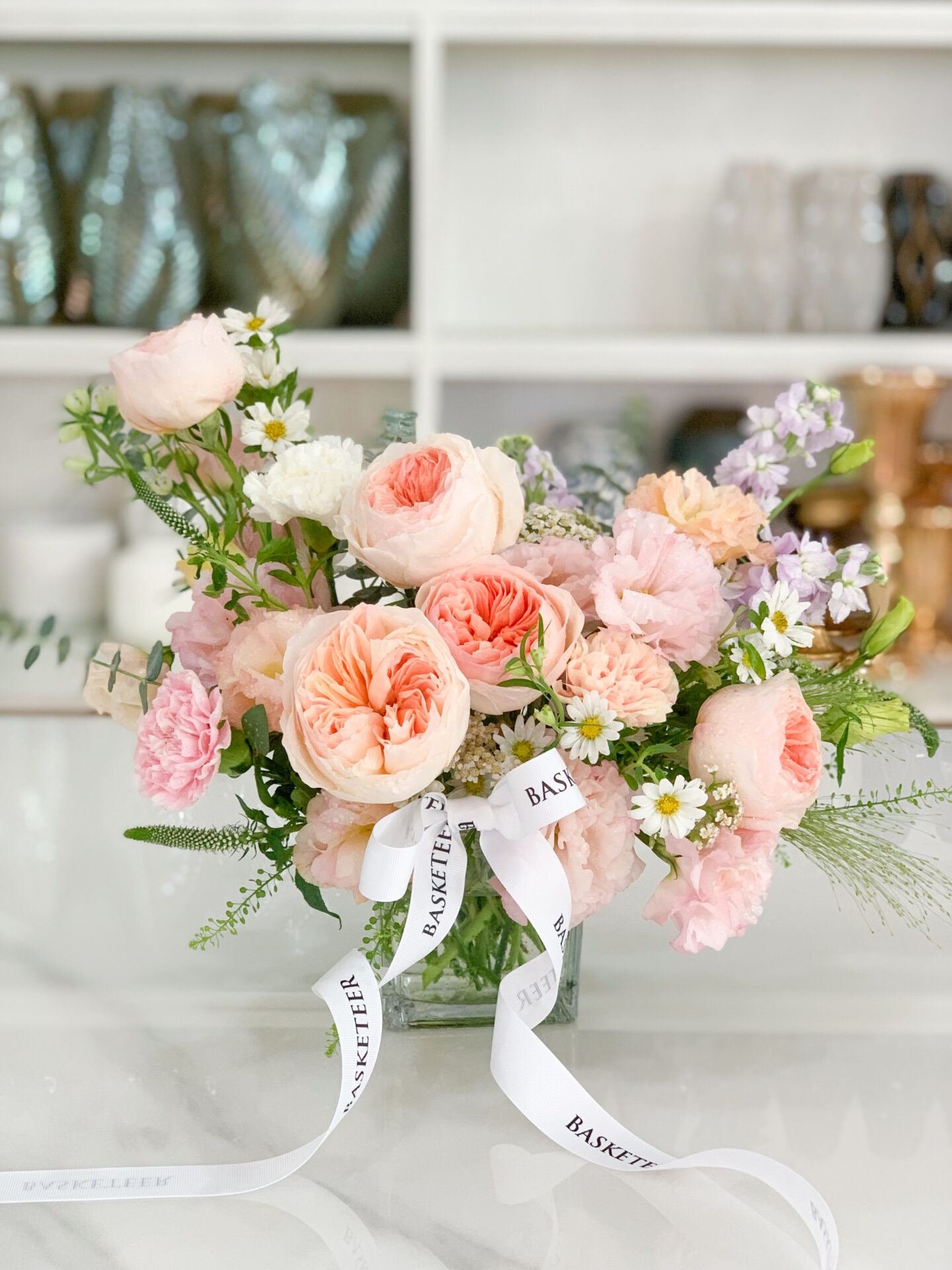 A flower arrangement in a clear rectangular vase sits on a white surface. The bouquet includes peach and pink roses, white daisies with yellow centers, and green foliage. White ribbons with the text 