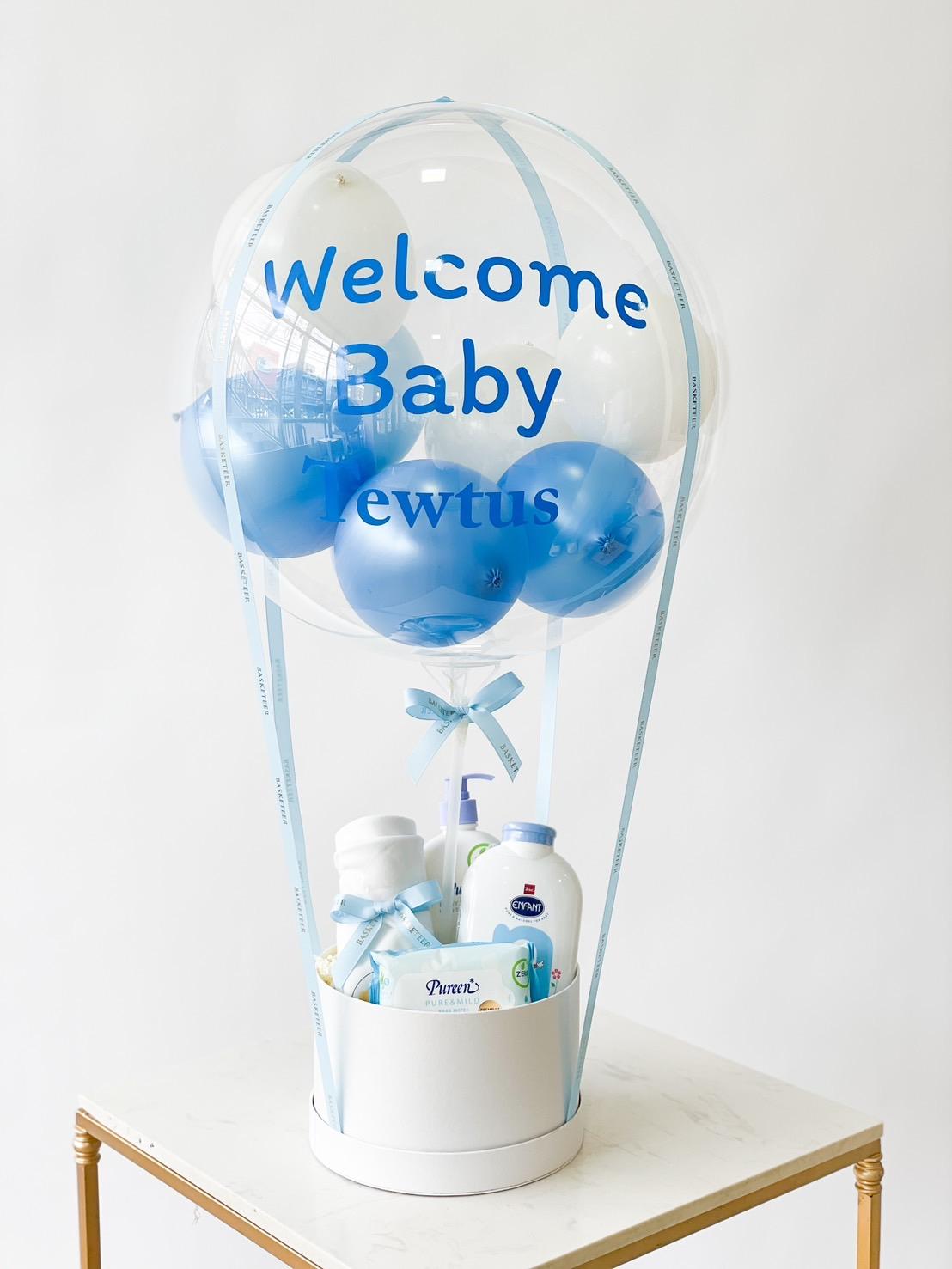 A baby gift arrangement resembling a hot air balloon includes a clear balloon with blue and white smaller balloons inside and the words 