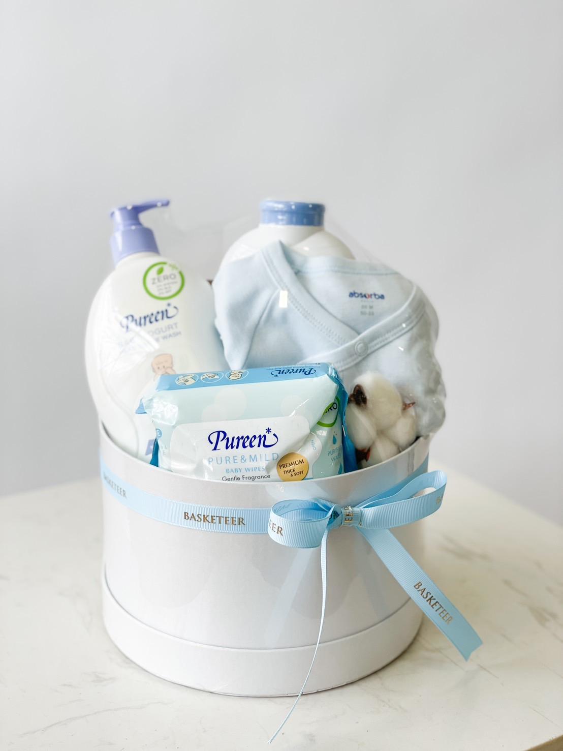 The Sweet Dreams Newborn Arrival Set includes a white gift basket with a blue ribbon, containing baby essentials: a bottle of Pureen baby lotion, a pack of Pureen gentle baby wipes, and folded baby clothes including a blue onesie, all neatly arranged against a light grey background.