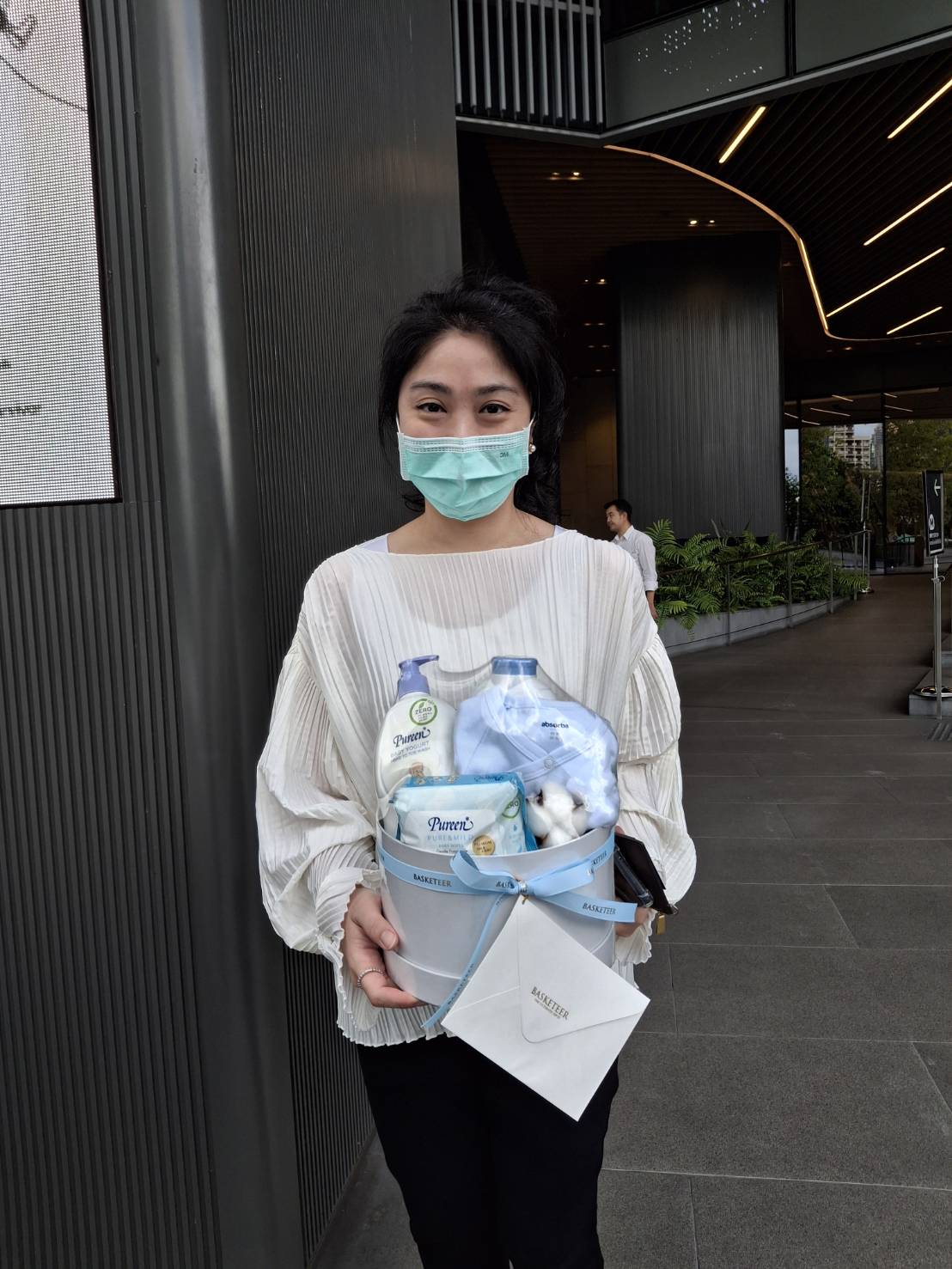 A woman wearing a face mask holds a festive gift basket, including the Sweet Dreams Newborn Arrival Set, with assorted items like a bottle of lotion. She stands in front of a modern building entrance, dressed in a white blouse and black pants.