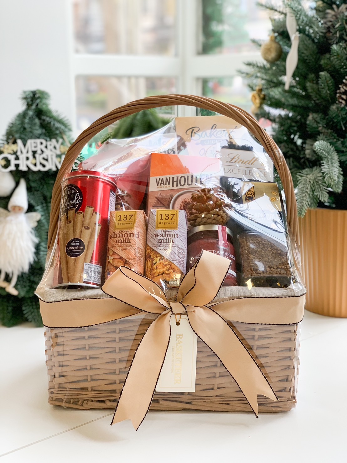 A wicker gift basket filled with almond and walnut milk, coffee, chocolate wafers, and snacks, tied with a beige ribbon.