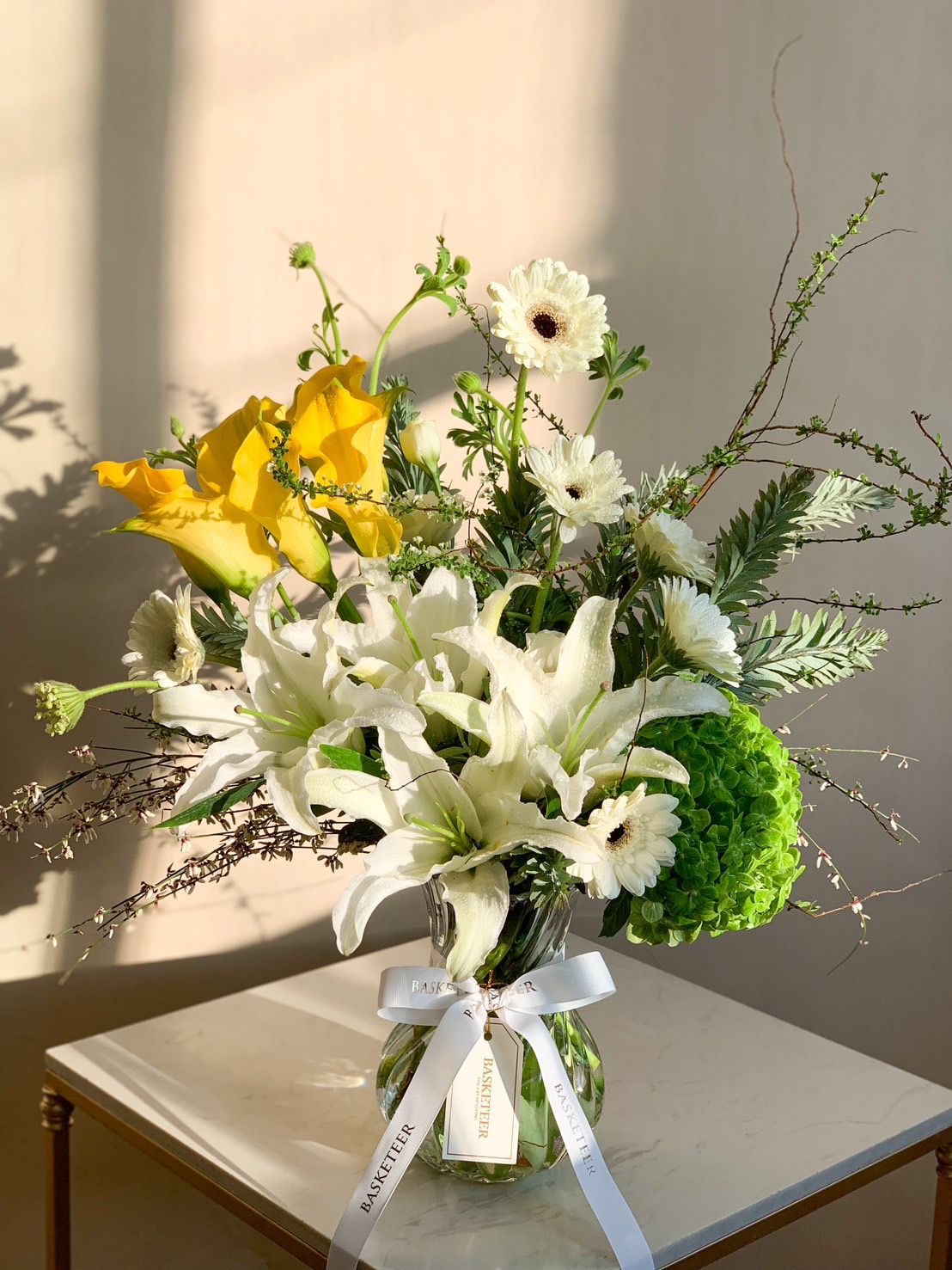 A vibrant floral arrangement in a glass vase featuring yellow calla lilies, white lilies, and hydrangeas, decorated with a white ribbon.