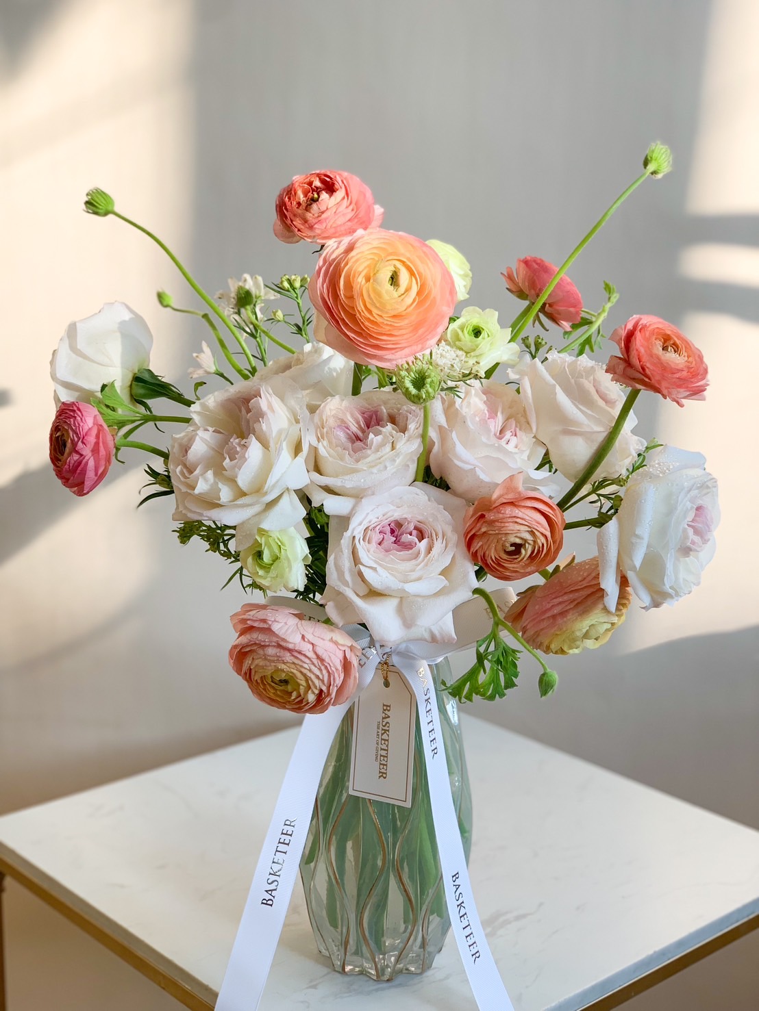 A beautiful bouquet of pastel ranunculus and roses in shades of blush, peach, and white, arranged in a glass vase and tied with a white ribbon.