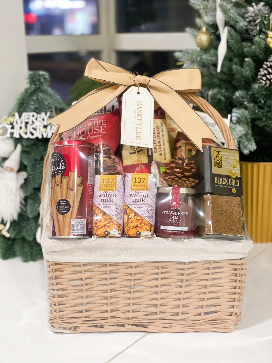 A woven gift basket filled with walnut milk, chocolate wafers, brownie bites, strawberry jam, and coffee, tied with a beige bow.