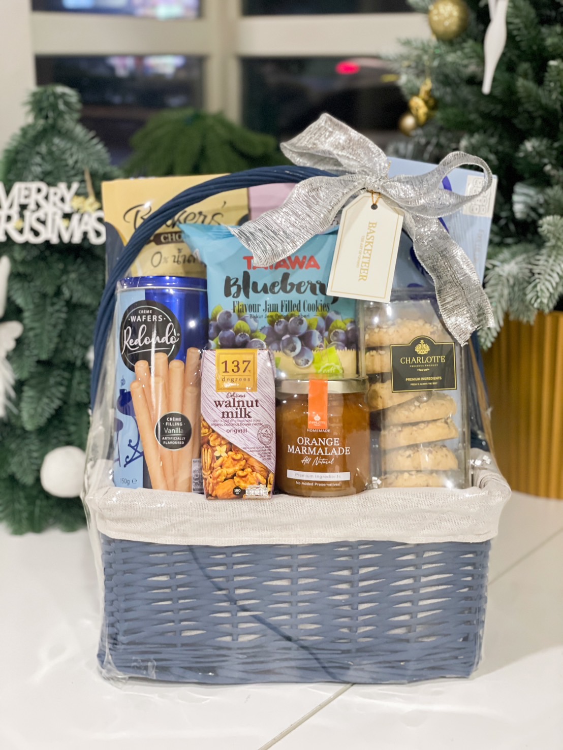 Blue basket filled with orange marmalade, almond walnut milk, chocolate wafers, blueberry jam cookies, and gourmet treats, tied with a silver bow.