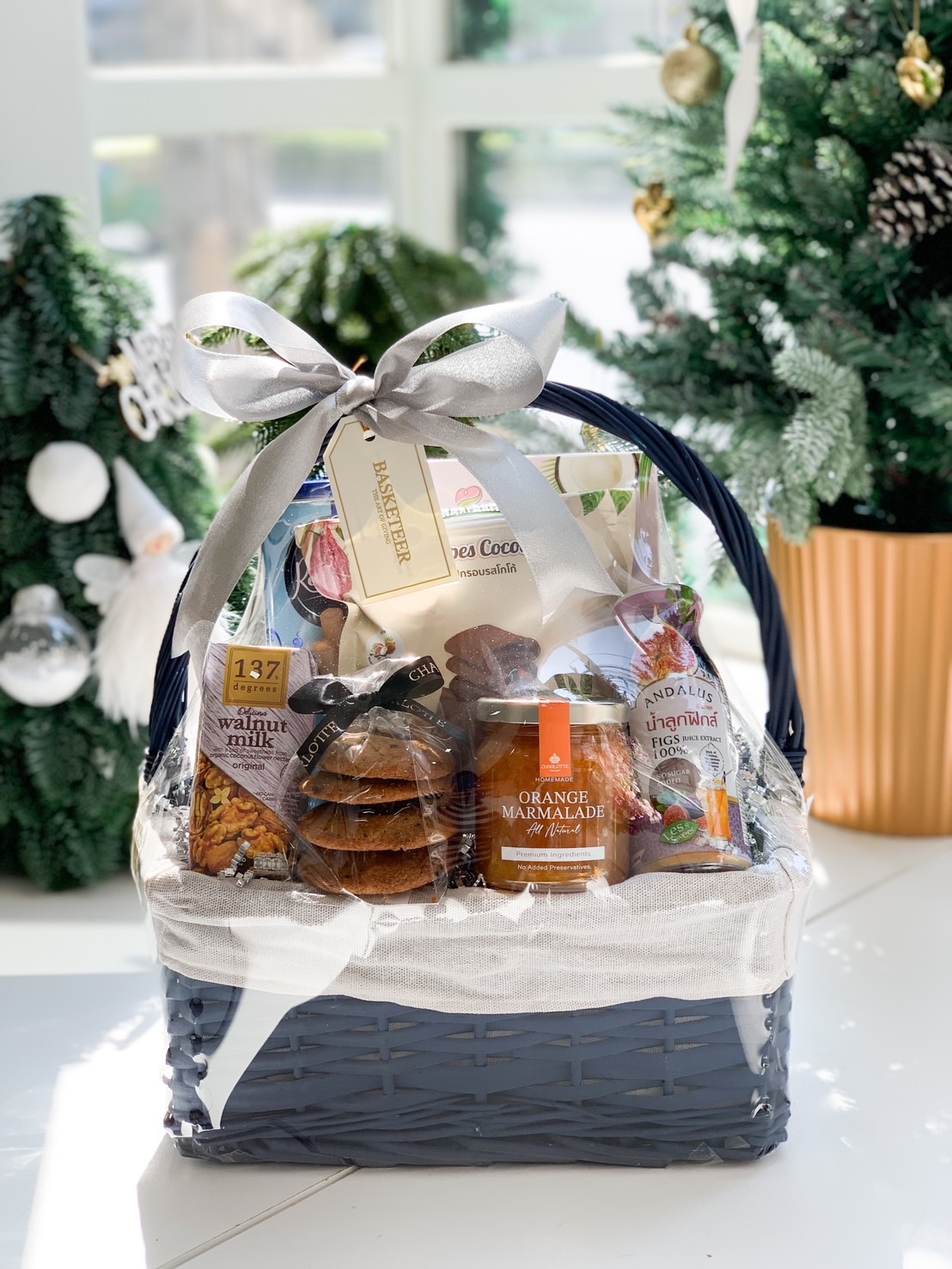 Dark blue gift basket filled with gourmet items including walnut milk, orange marmalade, cookies, and fig juice, tied with a silver ribbon and displayed near a festive Christmas tree.