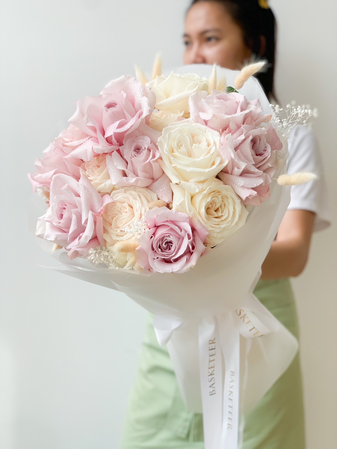 A person in a green apron holds a bouquet of white and pink roses wrapped in white paper. The Sweet Blooming Marvellous Roses Bouquet includes a ribbon with the word 