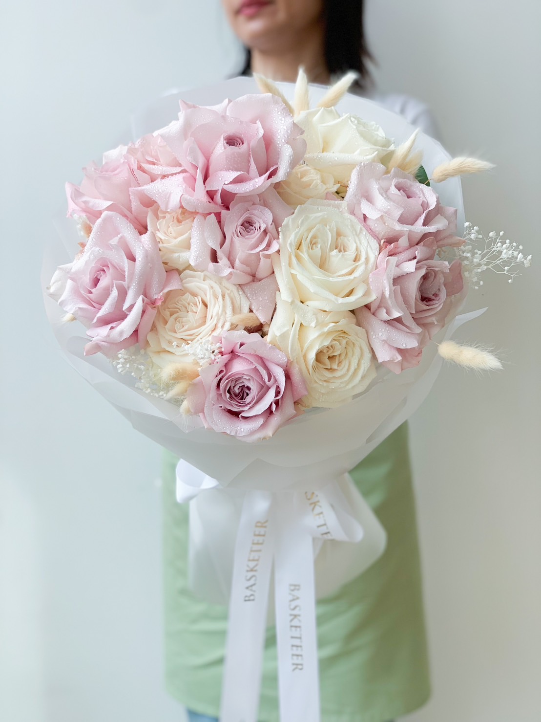 A person holding a Sweet Blooming Marvellous Roses Bouquet adorned with light pink and white roses, small white flowers, and green leaves. The bouquet is wrapped in white paper with ribbons that have 