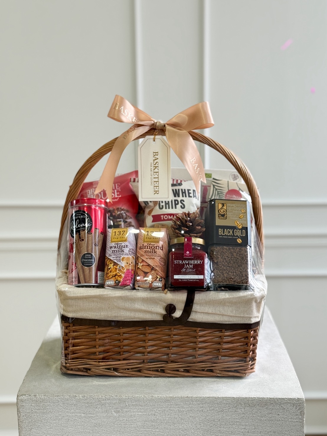 Luxurious Thai wicker gift hamper, elegantly wrapped, with gourmet snacks, premium drinks, chocolate, coffee, and a Basketier tag.