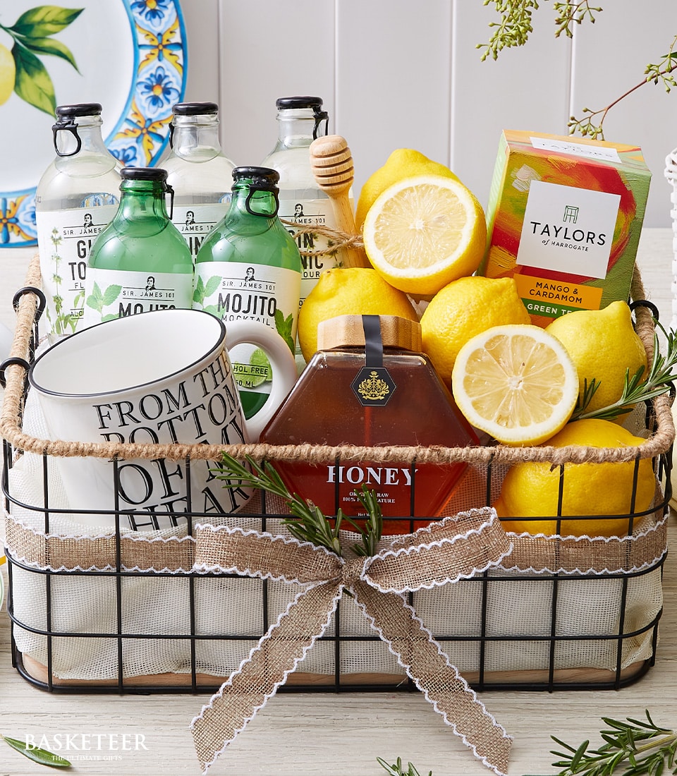 A luxurious basket adorned with the Basketeer logo, brimming with elegant delights: premium bottles of tonic water and exquisite mojito mix, a jar of golden honey paired with a handcrafted honey dipper, three vibrant lemons for that perfect zest, an upscale box of Taylors tea, two pristine white mugs ready for a comforting brew, a radiant yellow hassock flower adding a touch of charm, and fragrant rosemary sprigs to elevate any experience.