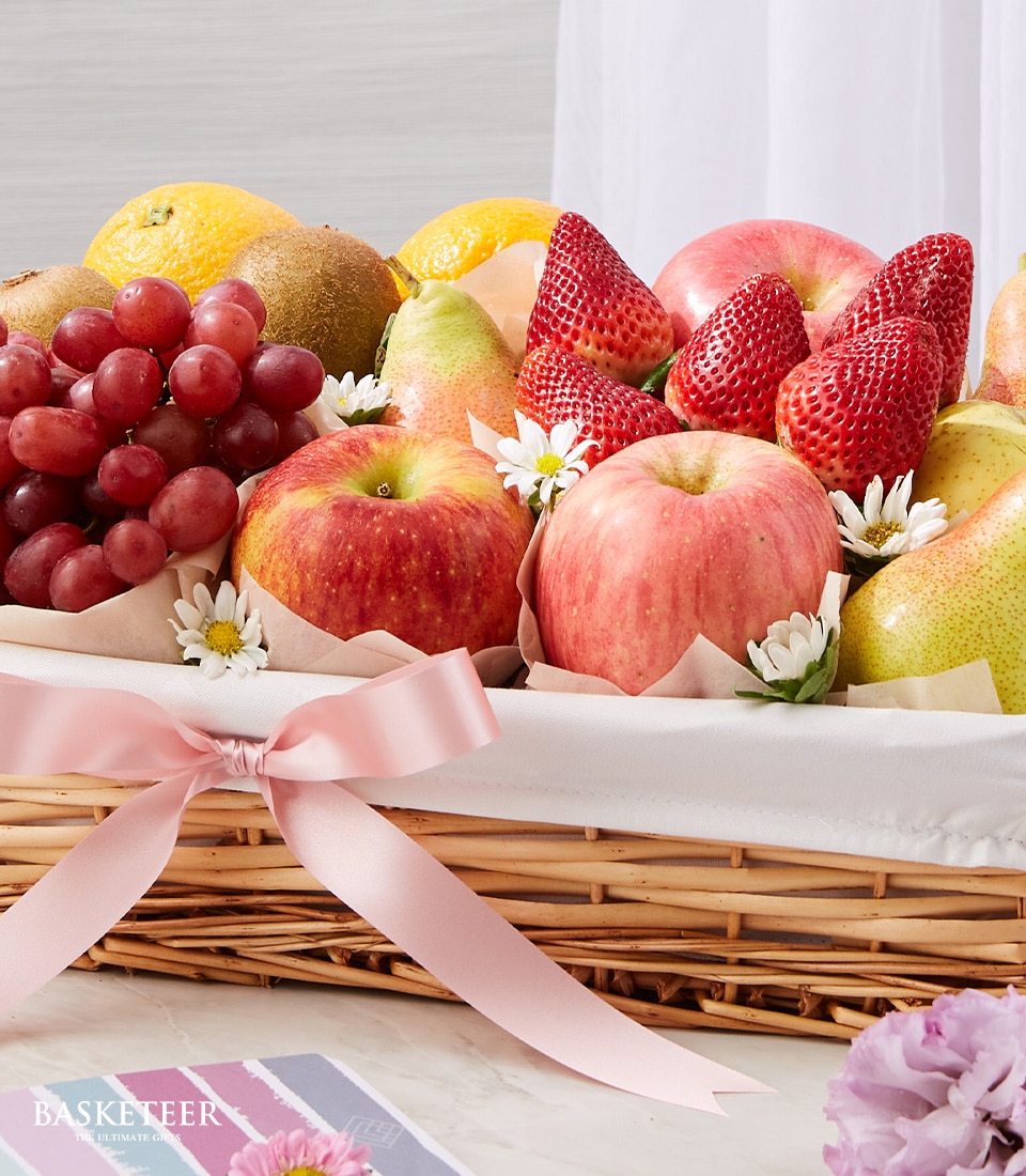 A lavish Basketeer Fruit Abundance Basket overflowing with an assortment of the freshest fruits, including crisp apples, succulent strawberries, juicy grapes, and vibrant oranges. The exquisite arrangement is beautifully garnished with delicate white and yellow blossoms. The basket features an elegant pink ribbon bow and a pristine white cloth lining, creating an inviting and luxurious presentation. An emblematic touch of Basketeer branding enhances this magnificent fruit display.

