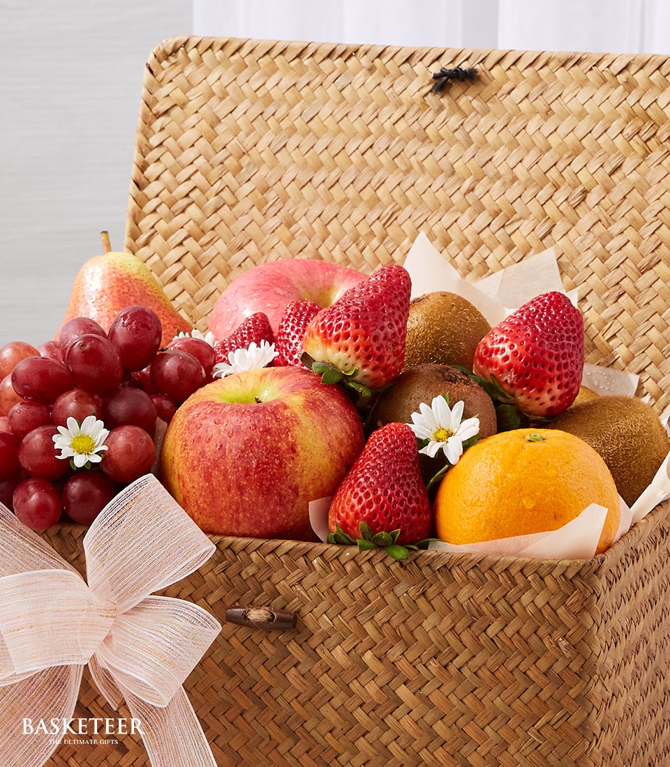 A luxurious Gourmet Fruit Celebrations Basket by Basketeer, elegantly showcasing a carefully curated selection of the finest fresh fruits. Indulge in the crisp perfection of red apples, the succulent ripeness of strawberries, juicy oranges, and tangy kiwis - all complemented by a lush bunch of ruby red grapes. This exquisite ensemble is nestled in an artisanal wicker basket adorned with delicate white blooms and finished with a refined light pink ribbon. Emblazoned with the prestigious Basketeer logo, this gift exudes opulence and invites delight at every glance.
