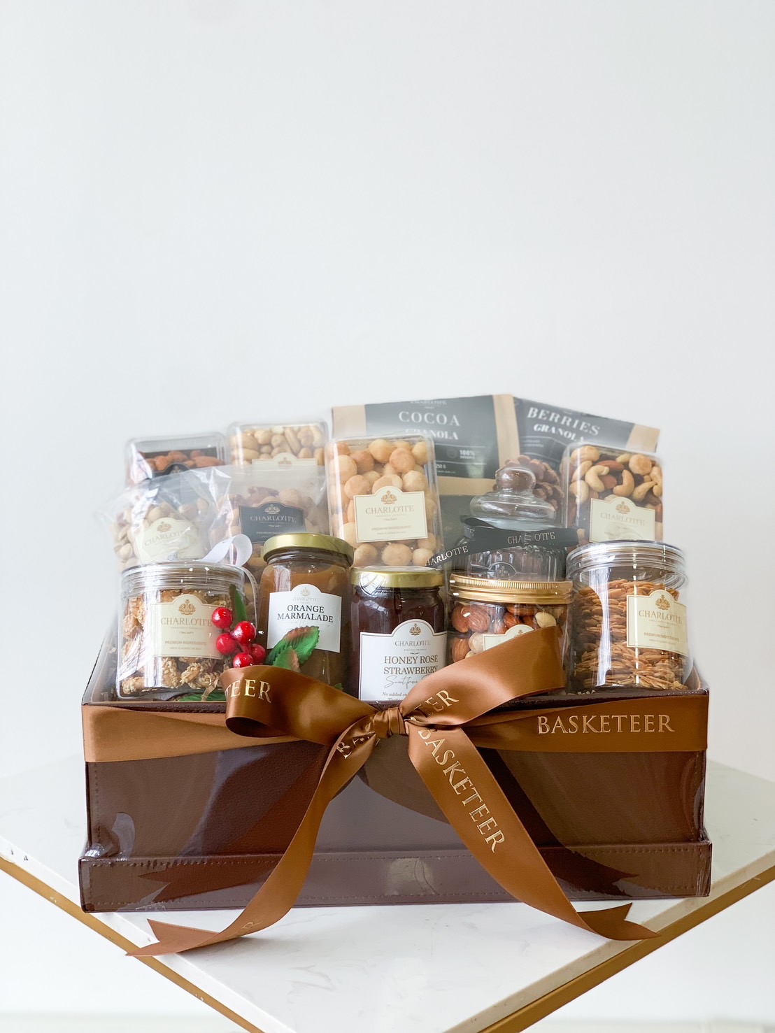 A gift basket featuring an assortment of nuts, dried fruits, and sweets in various jars and packets. The basket is adorned with a wide brown ribbon with the word 