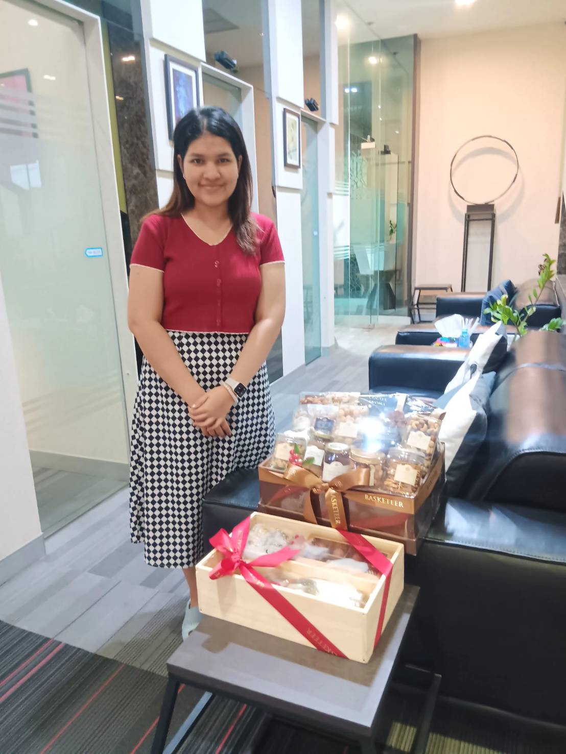 A woman with long hair stands in an office next to a table holding two gift boxes filled with various items. One box is decorated with a red ribbon. She is smiling and wearing a red top with a black and white checkered skirt. The background features modern office decor.