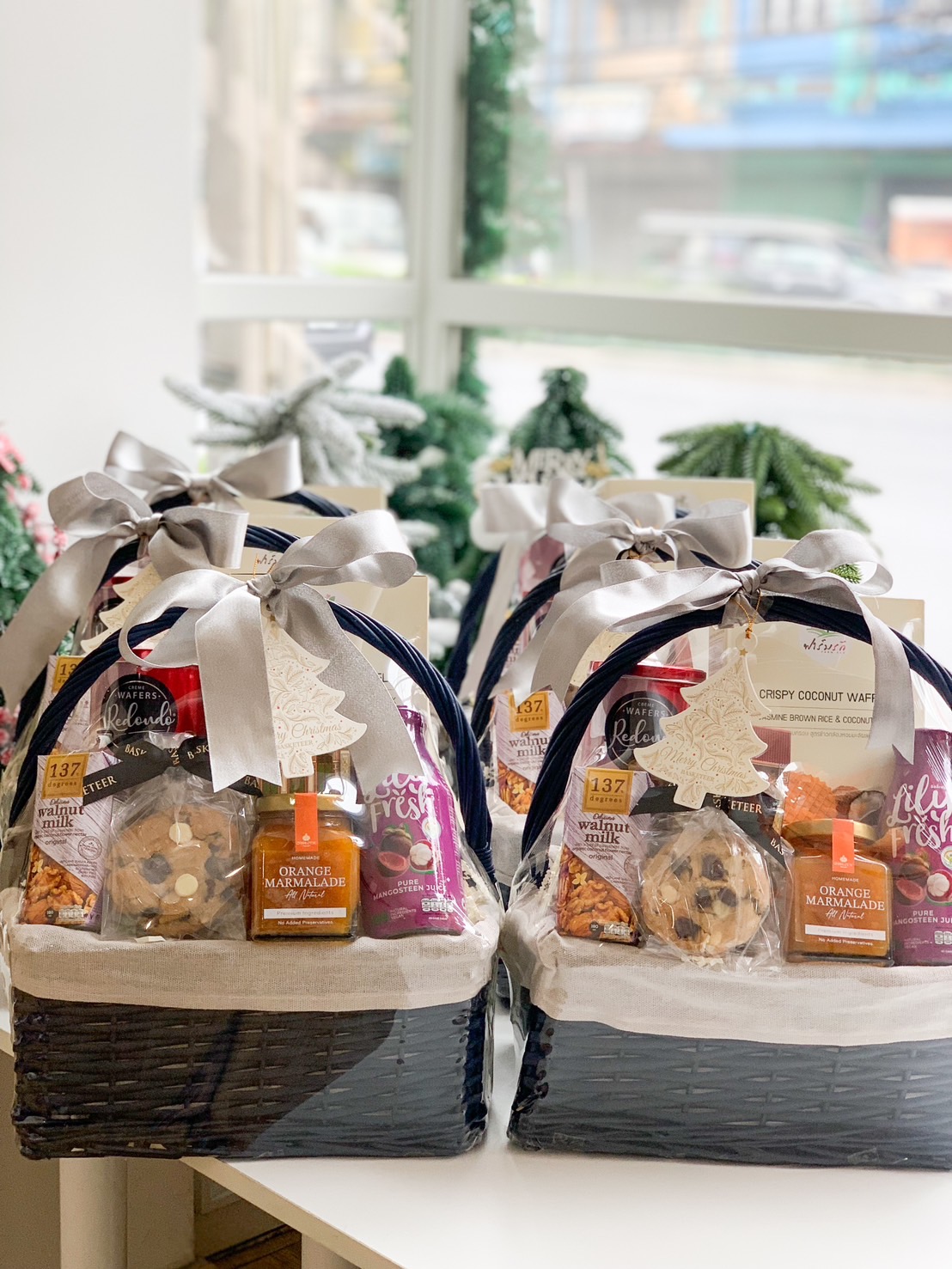 A lavish black wicker gift basket, elegantly lined with pristine white fabric, is presented by Basketeer. Inside, an array of gourmet delights awaits: melt-in-your-mouth chocolate chip cookies, a luscious Hershey's chocolate bar, delectable pineapple jam, indulgent crunchy almond crackers, and a refreshing can of fizzy drink. The ensemble is beautifully adorned with a sophisticated silver bow and proudly features the distinguished Basketeer logo on its ribbon.