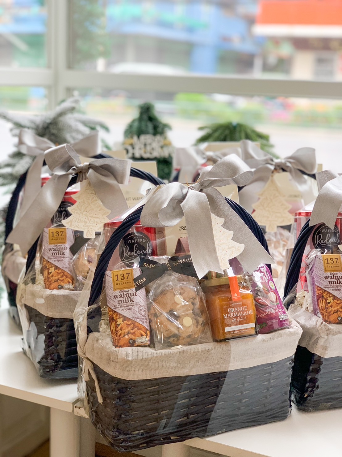 A lavish black wicker gift basket, elegantly lined with pristine white fabric, is presented by Basketeer. Inside, an array of gourmet delights awaits: melt-in-your-mouth chocolate chip cookies, a luscious Hershey's chocolate bar, delectable pineapple jam, indulgent crunchy almond crackers, and a refreshing can of fizzy drink. The ensemble is beautifully adorned with a sophisticated silver bow and proudly features the distinguished Basketeer logo on its ribbon.