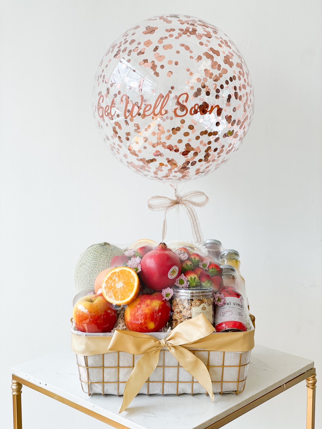 A Wellness Fresh Fruits Basket sits on a table, adorned with a large clear balloon with rose gold confetti and the words 