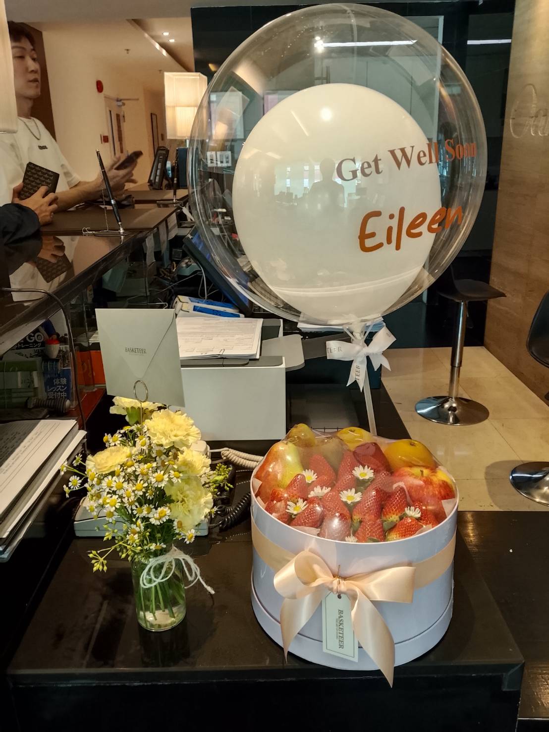 Get well soon gift set with a large clear balloon, fresh fruit box of strawberries, apples, pears, and small yellow flower vase.