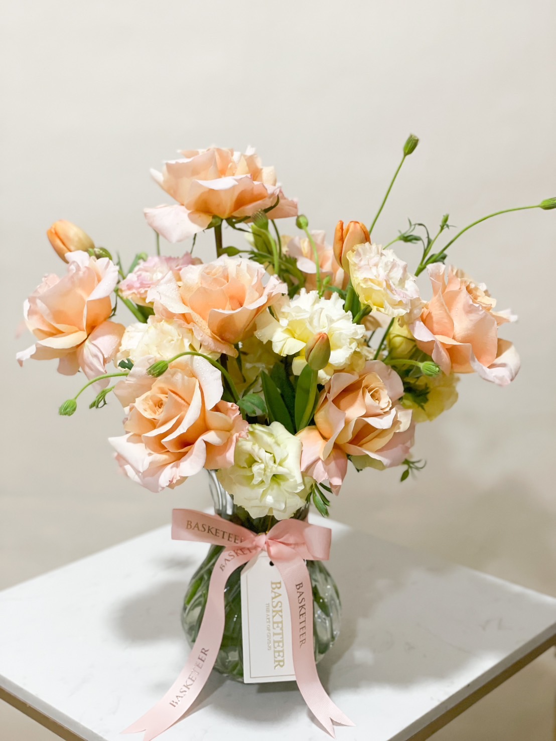 A luxurious peach and cream rose arrangement in a clear glass vase, adorned with lisianthus, delicate greenery, and tied with a soft pink ribbon.