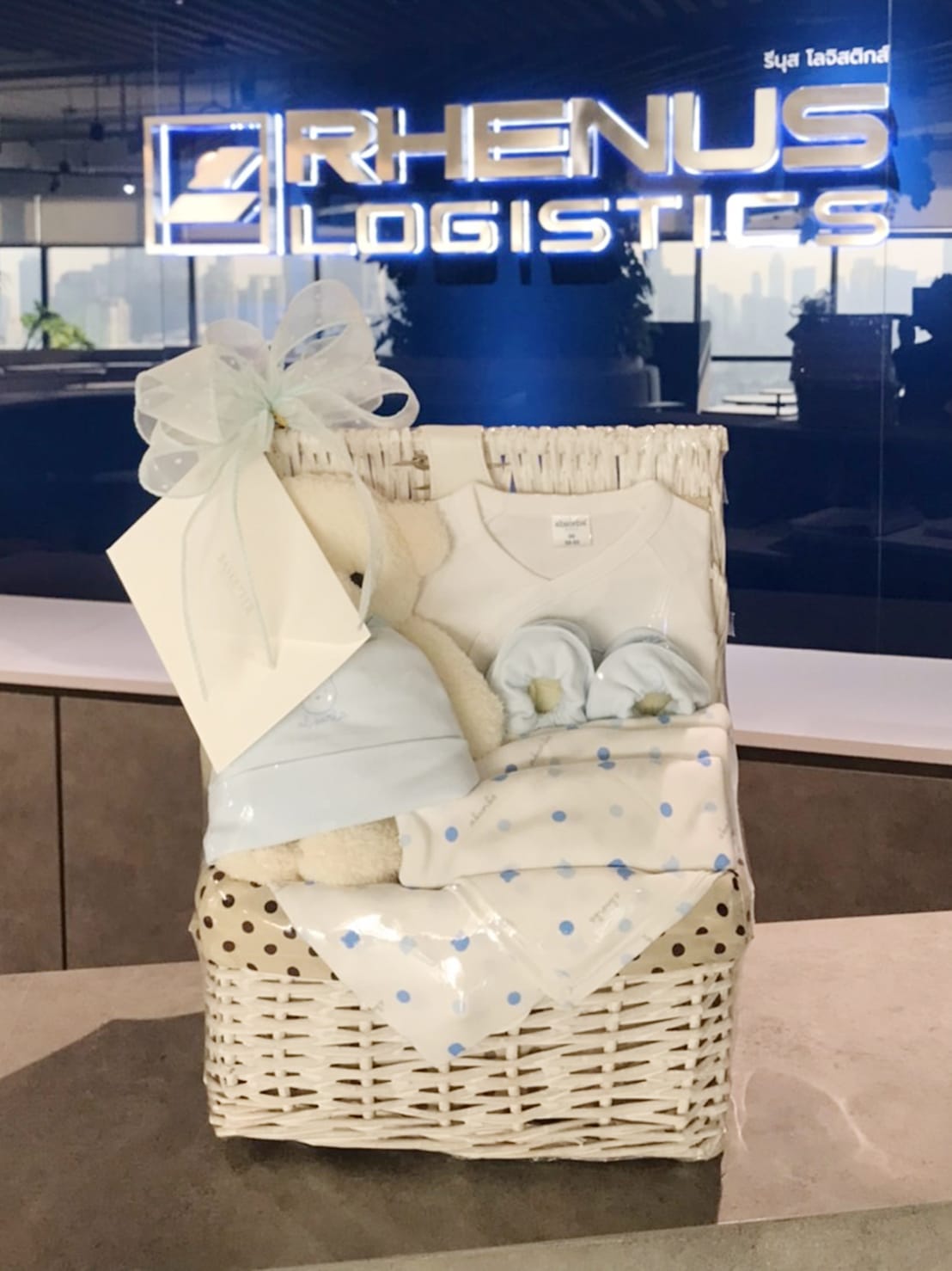 A white wicker gift basket contains a teddy bear, a baby onesie, a pair of baby booties, and a polka dot blanket, all in pastel blue and white. The basket is adorned with a blue ribbon and sits on a counter. The background features a Rhenus Logistics sign.