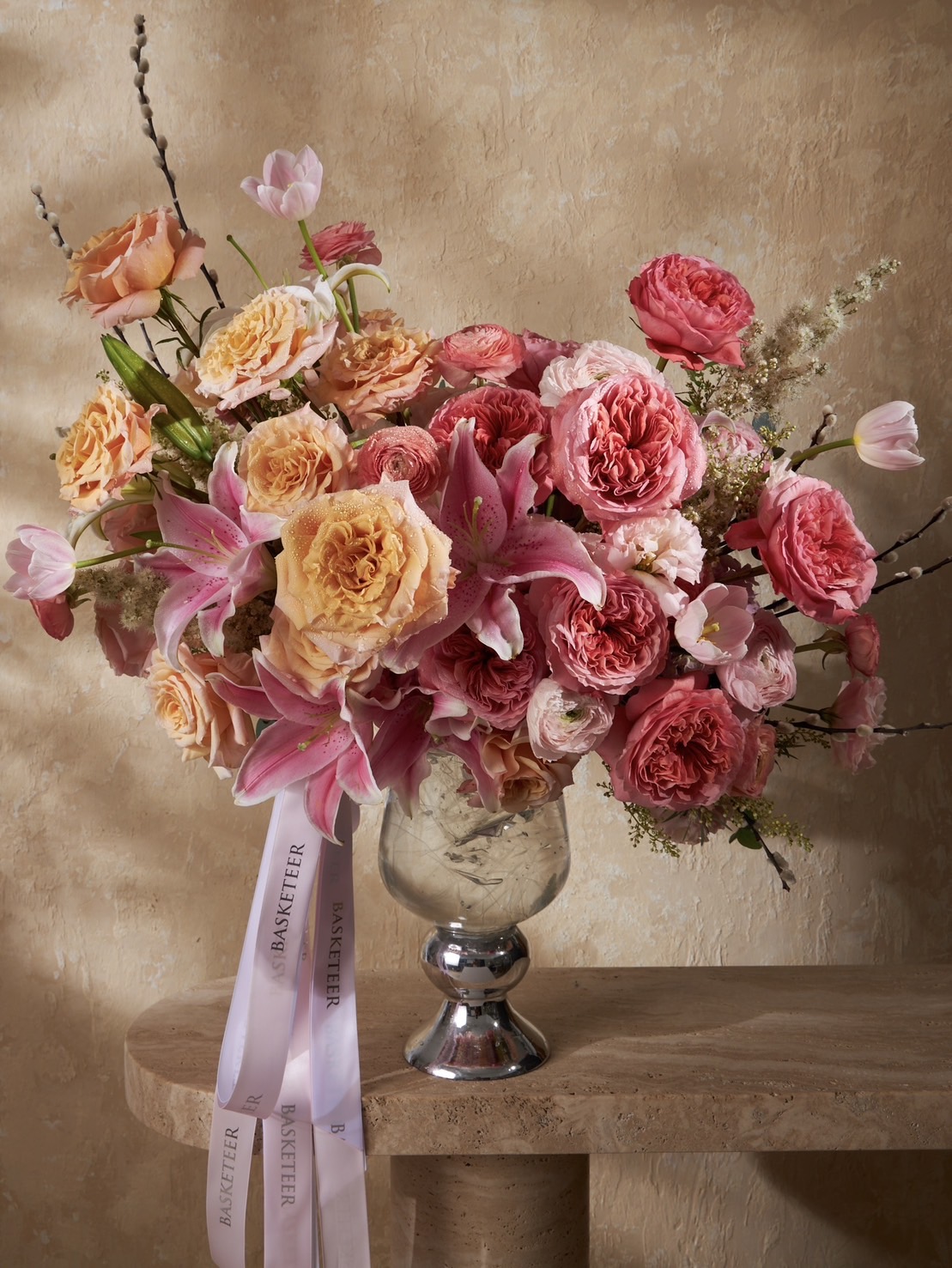 Elegant glass vase filled with peach roses, pink garden roses, lilies, and tulips in a romantic pink-peach arrangement with BASKETEER ribbons.
