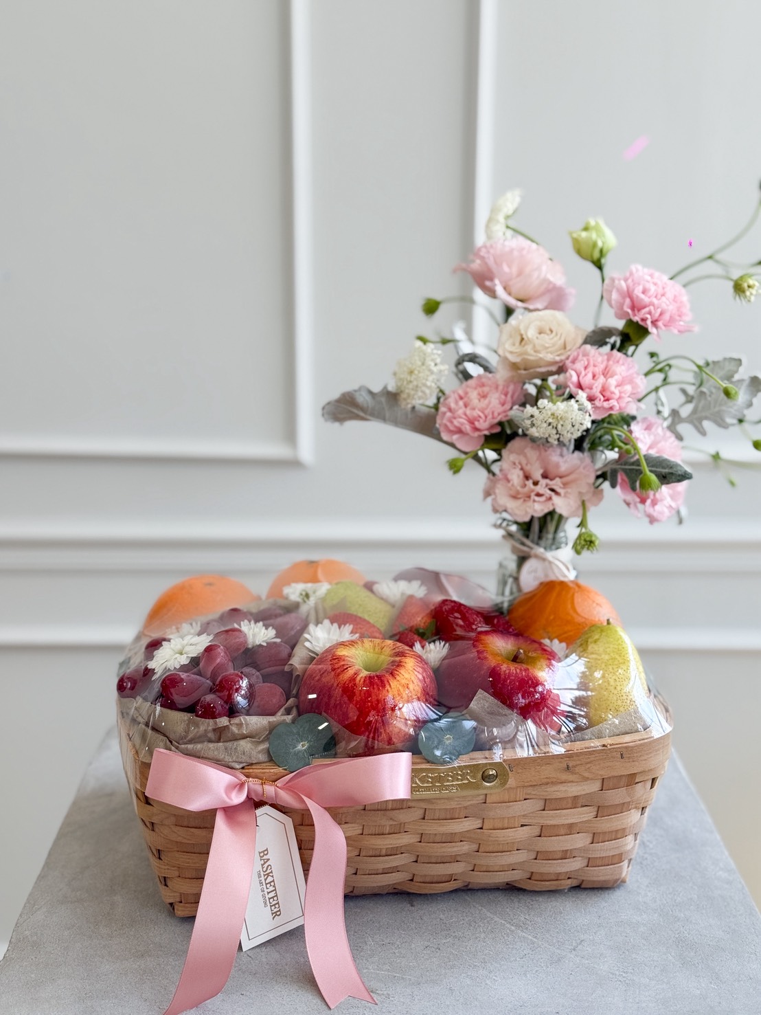 Elegant fruit basket with red apples, oranges, pears, grapes, and melon, topped with pink flowers and tied with a pink satin ribbon.