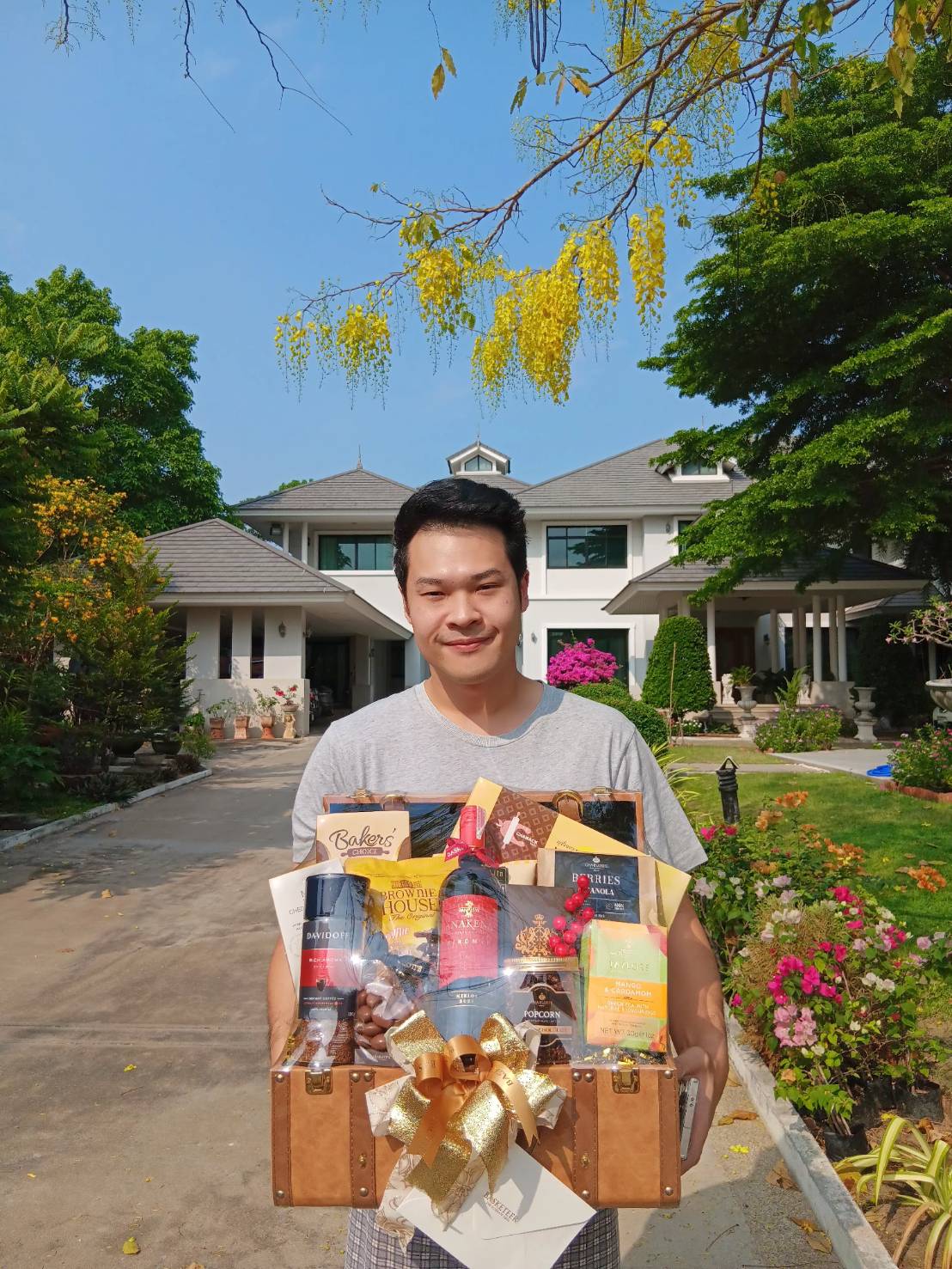 A smiling man in a gray t-shirt holds a gift basket filled with various items, including wine bottles, chocolates, and packaged goods. He stands outdoors on a sunny day, with a large house, trees, and colorful flowers in the background.