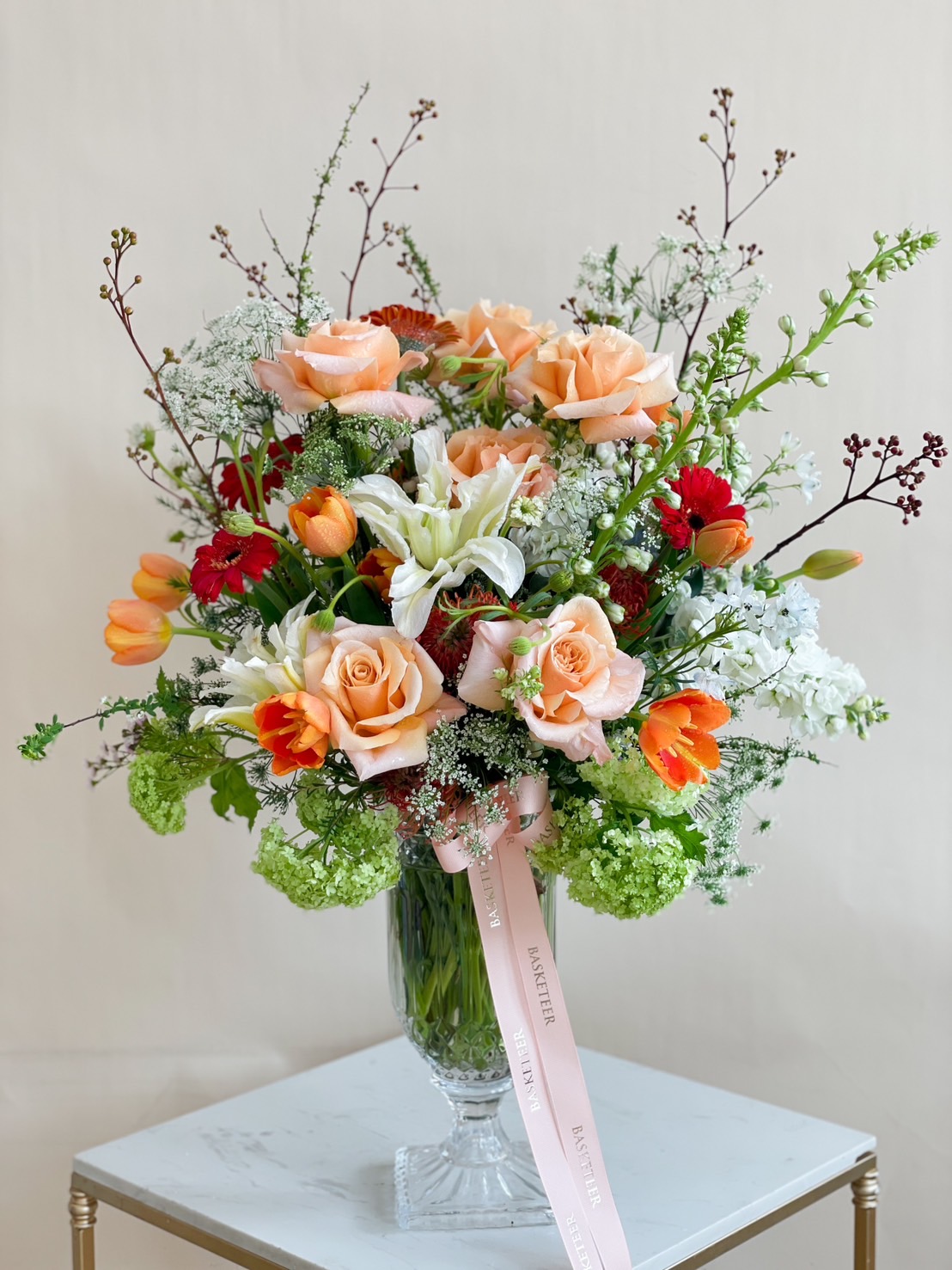 A premium fresh flower arrangement with peach roses, white lilies, tulips, and lush greenery in a crystal glass vase, ideal for luxury gifting and décor.