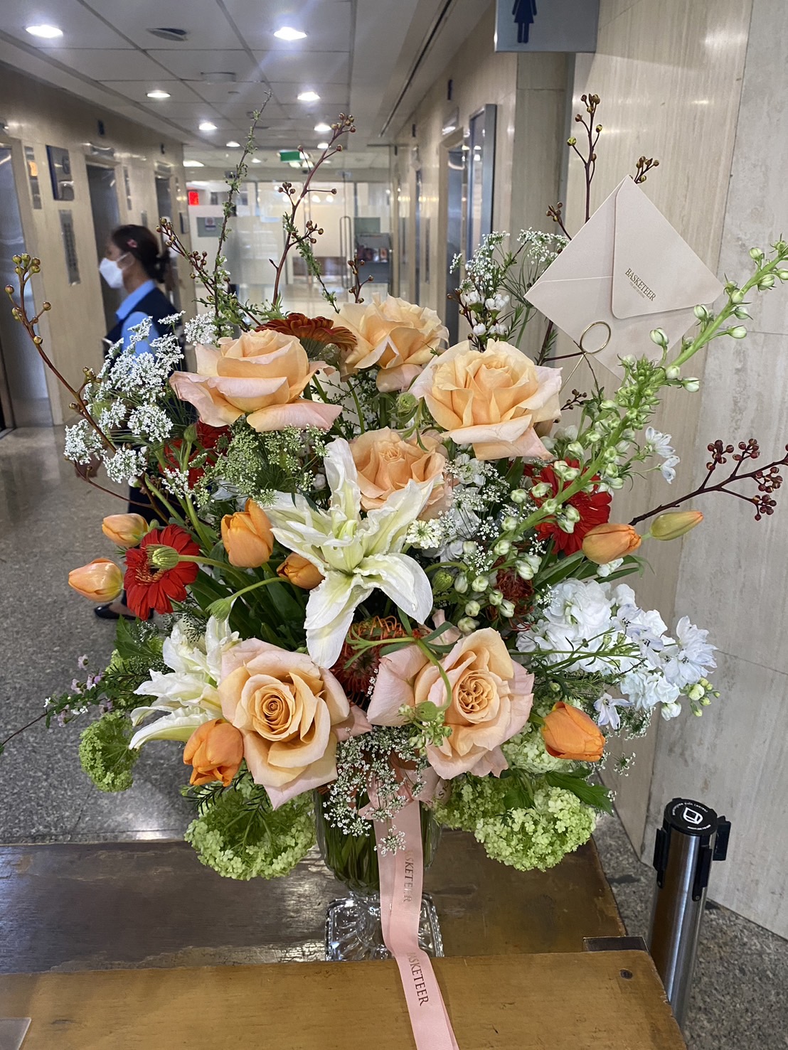 A stunning fresh flower arrangement with peach roses, white lilies, orange tulips, red gerberas, and lush greenery in a glass vase, ideal for gifting and décor.