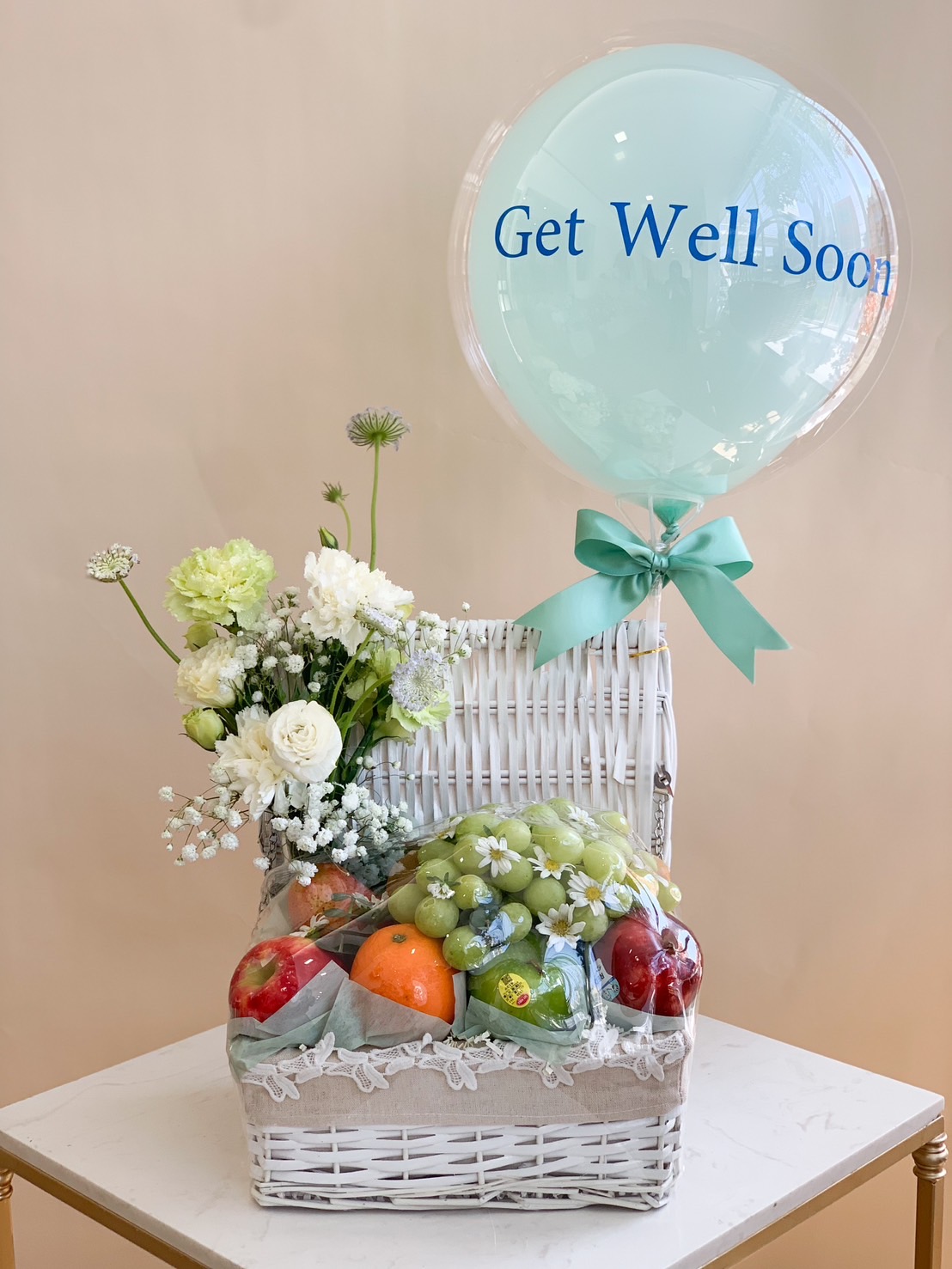 A beautifully arranged Get Well Soon fruit basket with fresh apples, grapes, and oranges, complemented by delicate white flowers and a pastel green balloon for a thoughtful recovery gift.