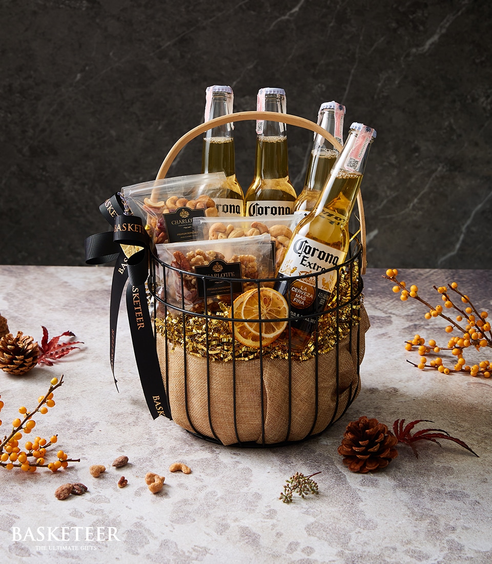 The opulent Beer Nut Haven Basket from Basketeer exudes luxury, featuring four Corona Extra bottles complemented by an exquisite selection of assorted nuts and dried fruits. The arrangement is adorned with shimmering golden tinsel, rustic burlap accents, and elegant black ribbons proudly showcasing the Basketeer logo. Nestled amidst artfully arranged pinecones and lush autumn foliage, this basket invites you to a rich sensory experience that combines craftsmanship with indulgence.