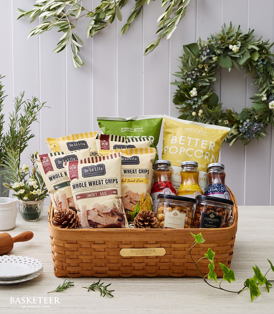 A luxurious wicker basket from Basketeer is brimming with an exquisite selection of snacks, including whole wheat chips, grilled corn chips, and perfectly popped popcorn. Nestled amongst the treats are elegant glass jars filled with golden honey, delectable jam, and gourmet mustard. The basket is artfully decorated with rustic pinecones and lush greenery. Positioned on a sophisticated table setting accented by a vibrant potted plant and a festive wreath in the background, this scene exudes warmth and refined charm. The signature Basketeer ribbon elegantly drapes across the basket’s handles, completing this inviting tableau of indulgence.