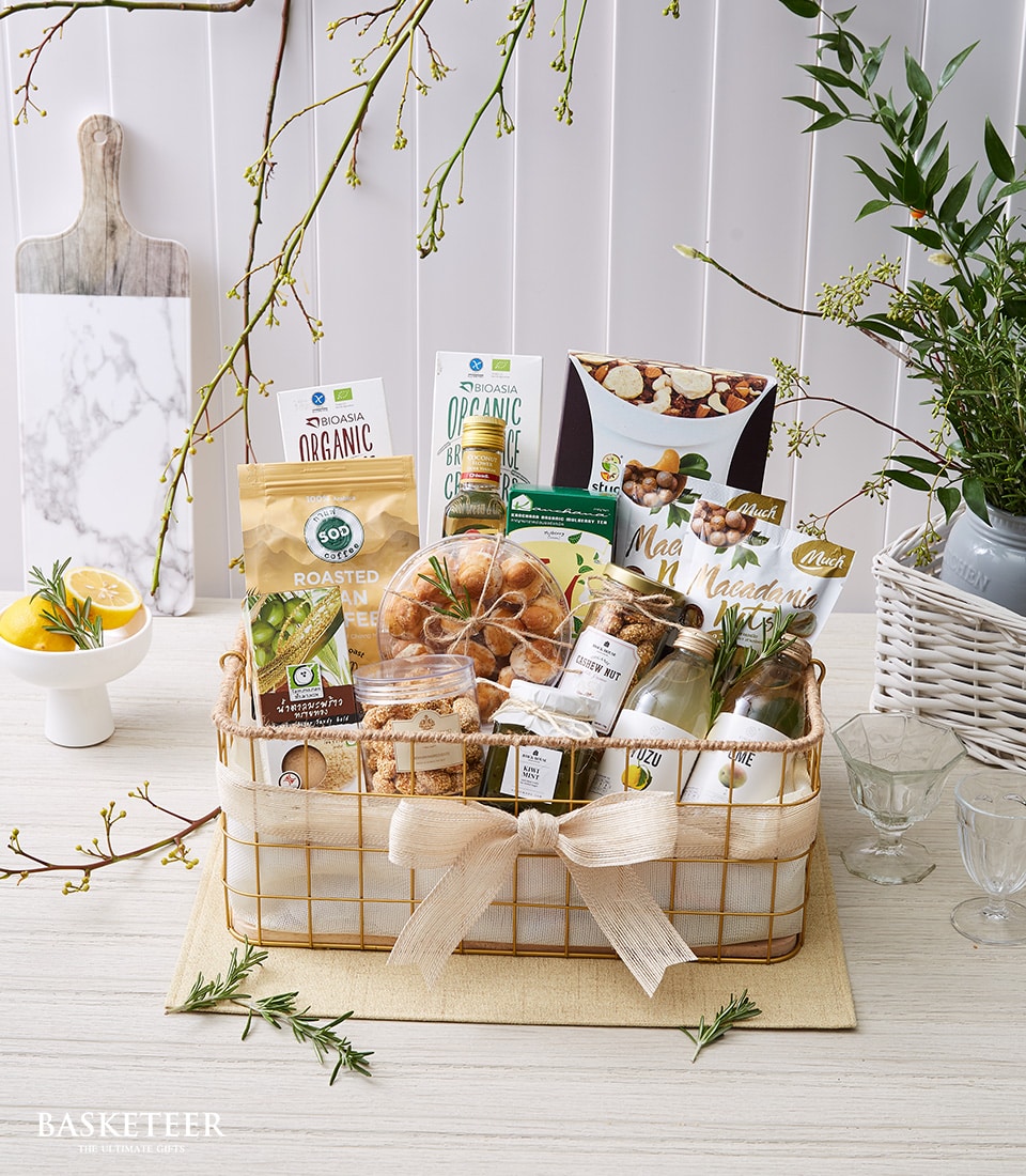 กระเช้าของขวัญสุดหรูที่เต็มไปด้วยของว่างและเครื่องดื่มออร์แกนิกคุณภาพเยี่ยมจาก Basketeer เพลิดเพลินกับเม็ดมะม่วงหิมพานต์ย่าง ถั่วรสเลิศหลากหลายชนิด ชาสมุนไพรชั้นเลิศ และขนมแสนอร่อยอื่น ๆ ประดับด้วยโบว์สีครีมหรูหราพร้อมโลโก้ Basketeer การจัดวางที่หรูหรานี้โดยมีฉากหลังเป็นต้นไม้เขียวขจีและมะนาวสีสันสดใสบนโต๊ะสีอ่อน เชิญชวนให้คุณดื่มด่ำกับความสง่างามอย่างแท้จริง