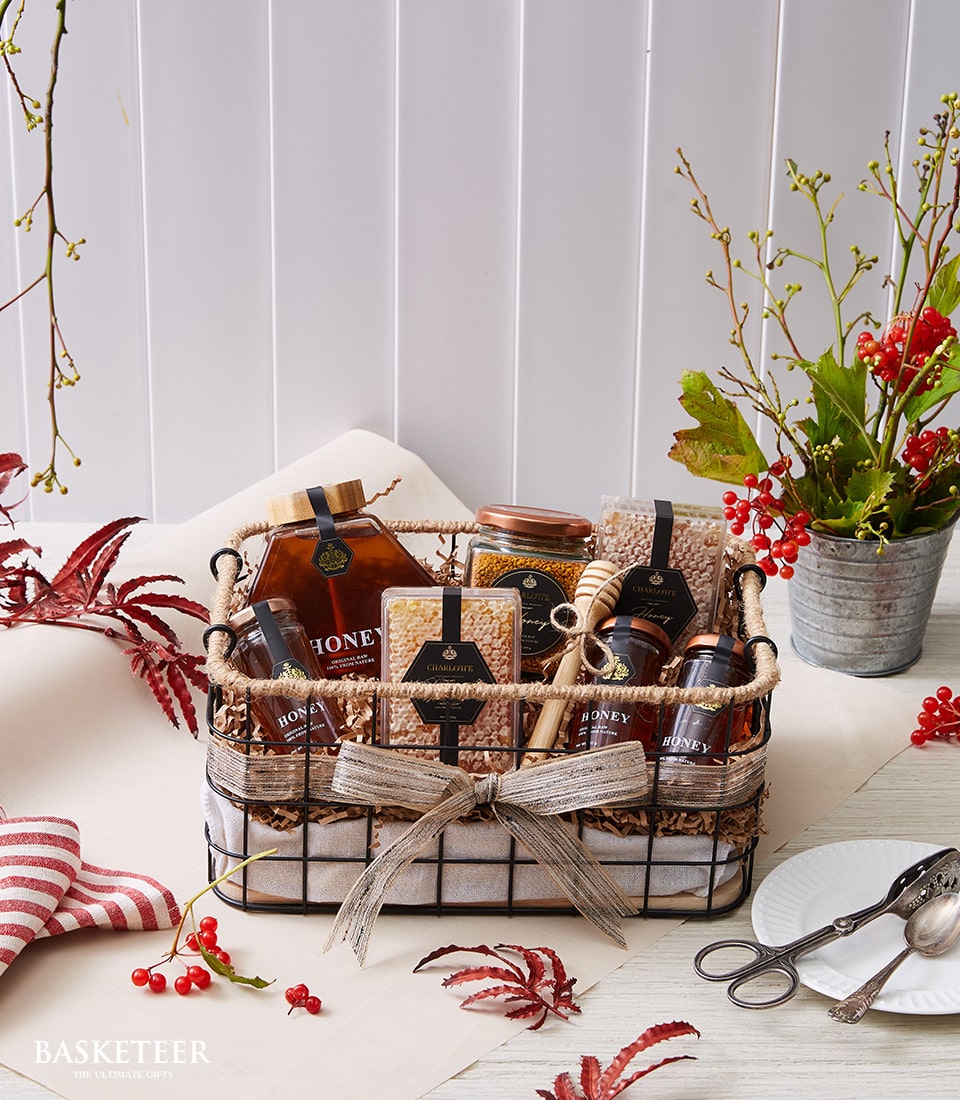 A lavish wicker basket, elegantly adorned with a satin bow, cradles an exquisite array of gourmet delights. Inside, you'll discover a premium bottle of honey, artisanal jars, fresh honeycomb, and beautifully wrapped goods. Each item promises indulgence and quality. The basket sits gracefully on a pristine white surface surrounded by seasonal accents—a potted plant with vibrant red berries, scattered autumn leaves, and additional meticulously wrapped packages that speak to the thoughtful curation of the Basketeer brand. Vertical blinds in the background accentuate the warm and inviting ambiance of this luxurious scene.
